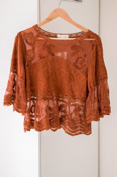 A rust-orange lace blouse with flared sleeves and intricate floral patterns hangs on a wooden hanger, perfect for any photography studio wardrobe, set against a clean white background.