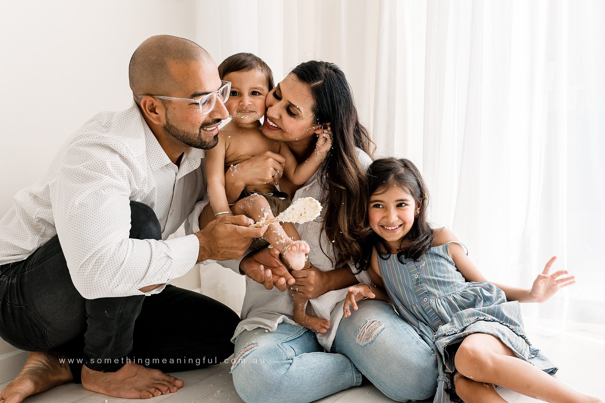 A joyful family of four sits together indoors, smiling and laughing. Captured by a Family and Newborn Photographer Melbourne West, they’re playfully covered in cake, with the father holding a spoon of frosting as light streams in through sheer curtains.