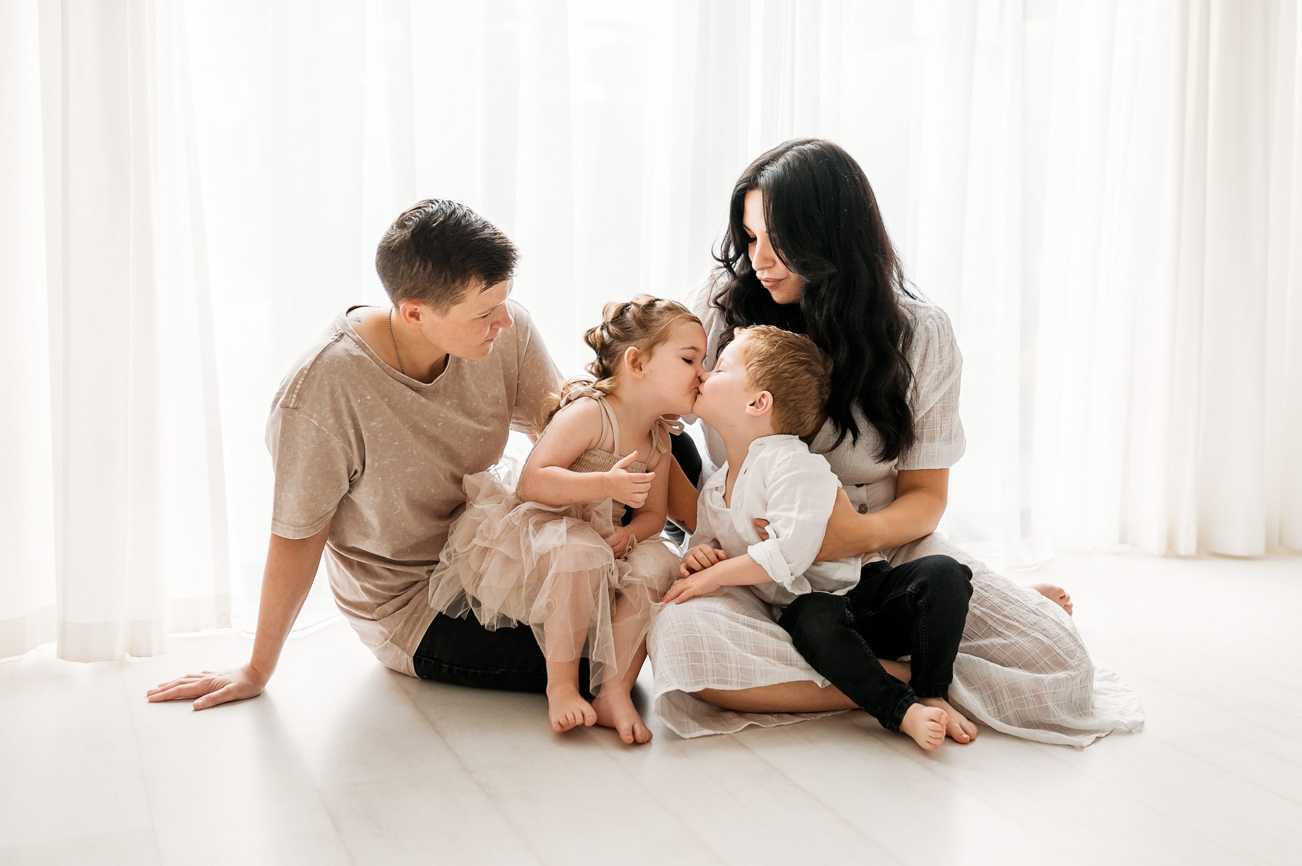 A family of four sits on the floor in a bright room. Two adults and two young children, one girl and one boy, share a tender moment as the children kiss each other—capturing the warmth of family photography Melbourne style. Everyone appears relaxed and happy.