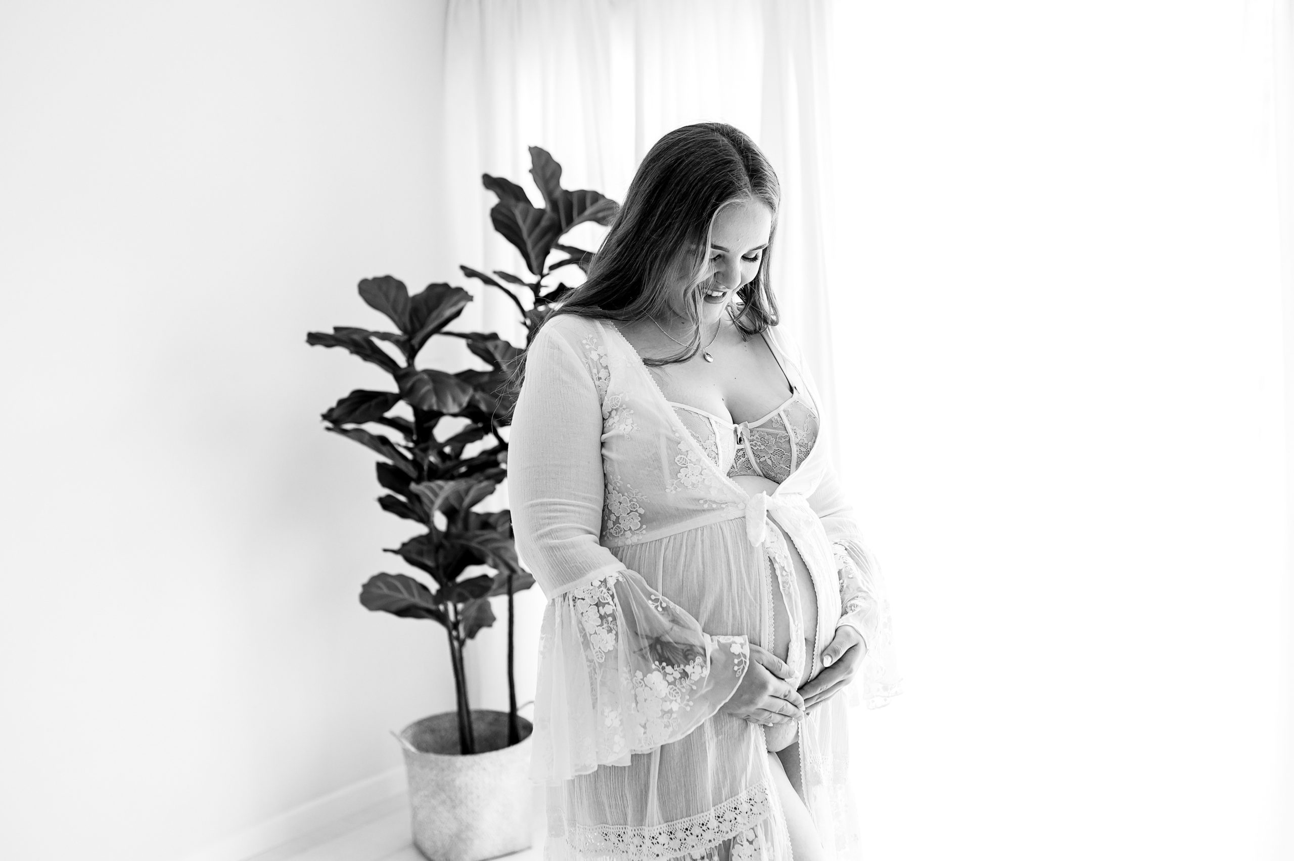 A pregnant woman in a lace robe stands indoors, gently holding her belly and smiling. A potted plant is beside her as soft natural light streams through sheer curtains—an elegant example of Maternity Photography Melbourne West. The image is in black and white.