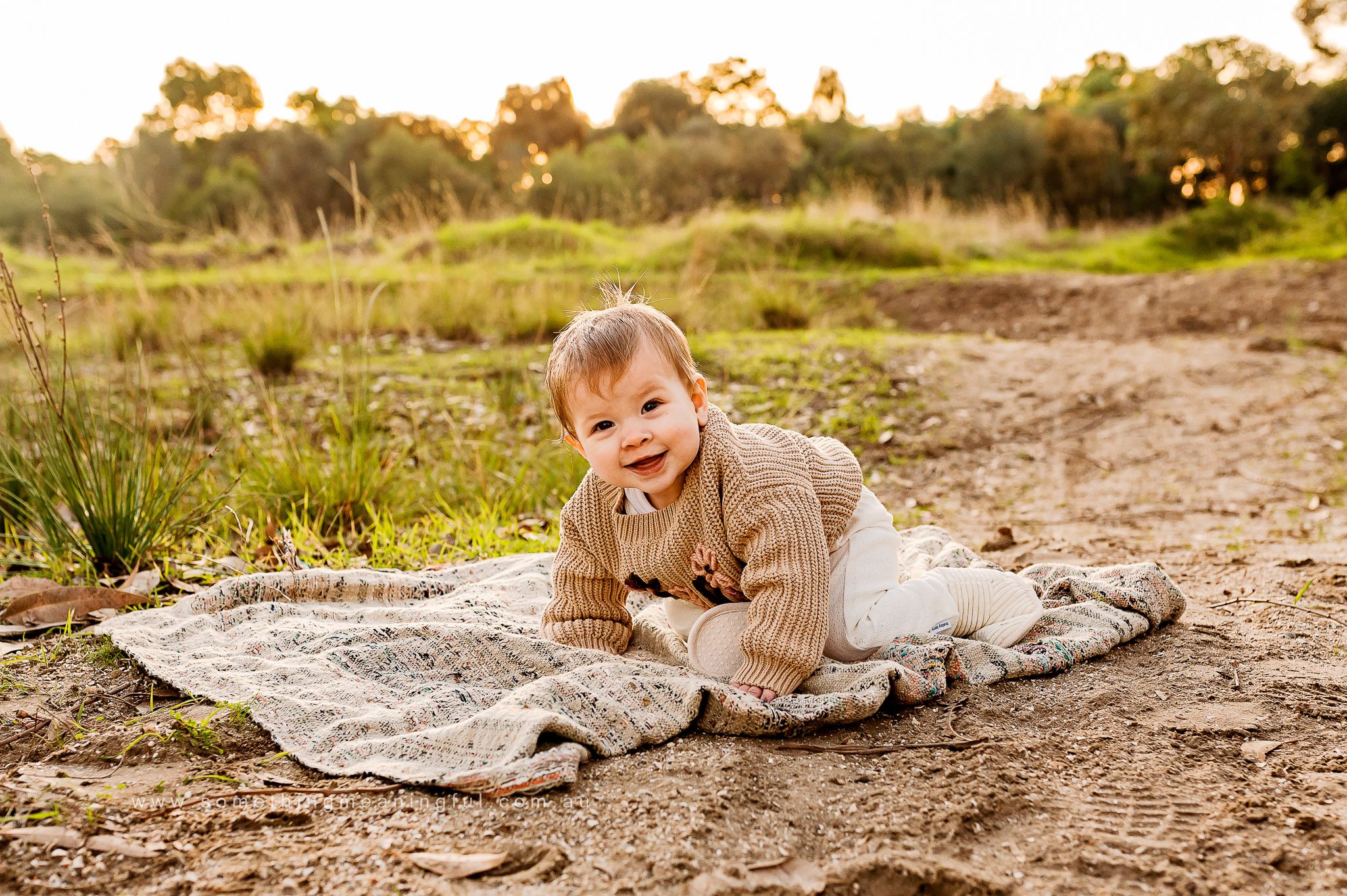 A smiling baby in a tan sweater and white pants crawls on a blanket outdoors, with green grass and trees glowing during golden hour—captured beautifully by a Melbourne family photographer.