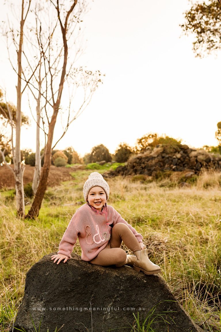 A young child wearing a knit hat, pink striped sweater, and boots sits smiling on a large rock in a grassy field with trees and golden sunlight in the background, captured by a Melbourne west family photographer.