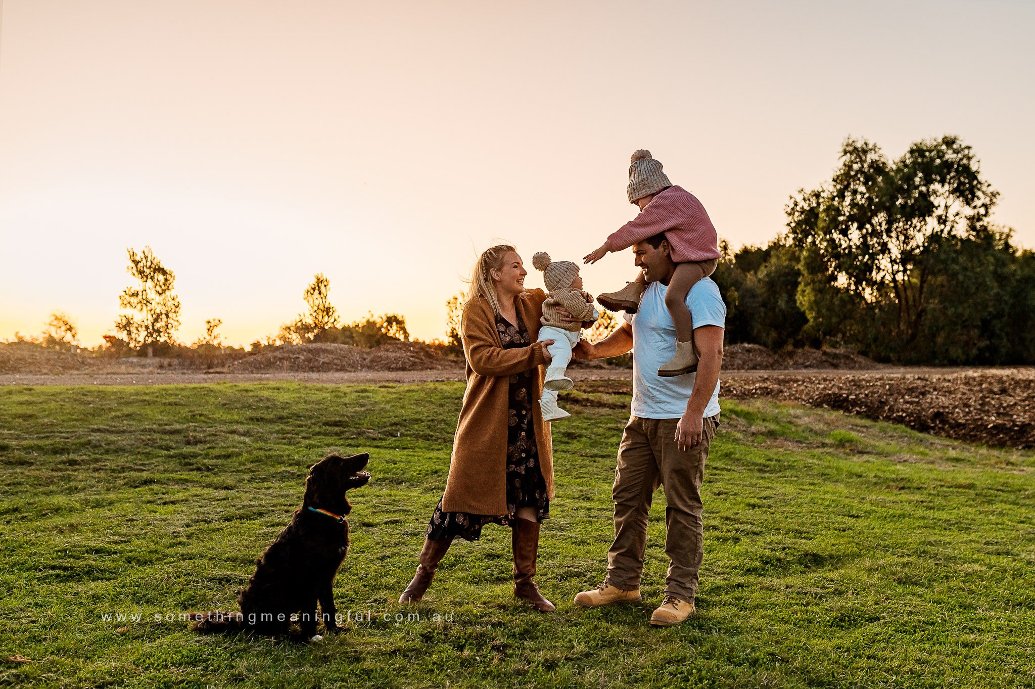 A family of four and their black dog stand on grass at sunset. Captured by a Melbourne family photographer, the mother holds a baby, the father lifts a child with a winter hat onto his shoulders. Trees and soft sunlight frame the scene.