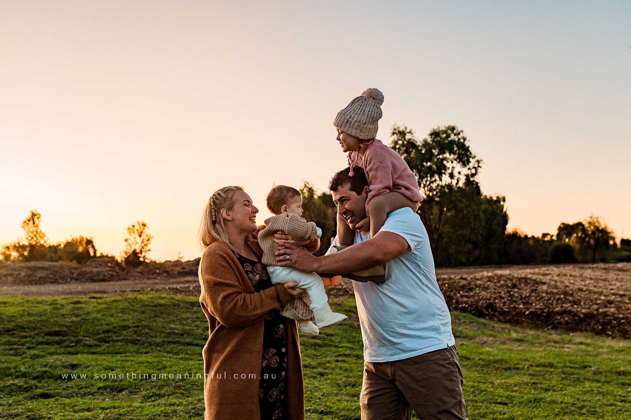 A family of four stands outdoors at sunset, smiling. Captured by a Melbourne family photographer, the mother holds a baby while the father carries a toddler on his shoulders. They appear happy, with trees and an open field in the background.