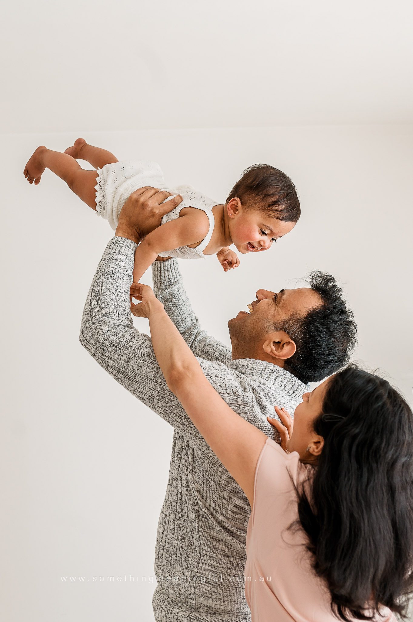 A man lifts a smiling baby in the air while a woman stands beside them, reaching towards the child. All three appear happy in a bright, minimal indoor setting—perfect for family photography with baby.
