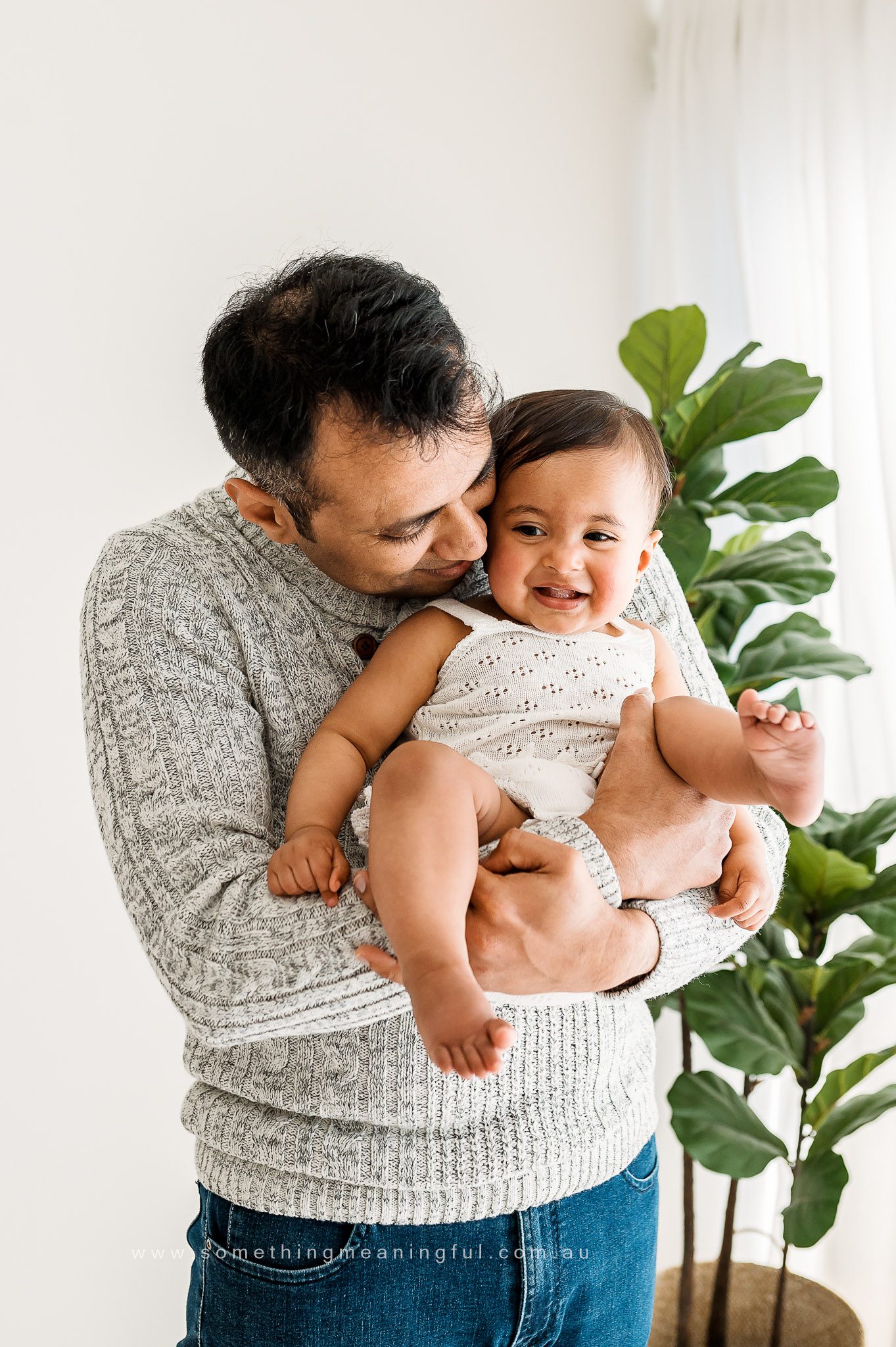 A man in a gray sweater lovingly holds and kisses a smiling baby dressed in white, capturing a tender moment of family photography with baby amid green leafy plants and white curtains in the background.