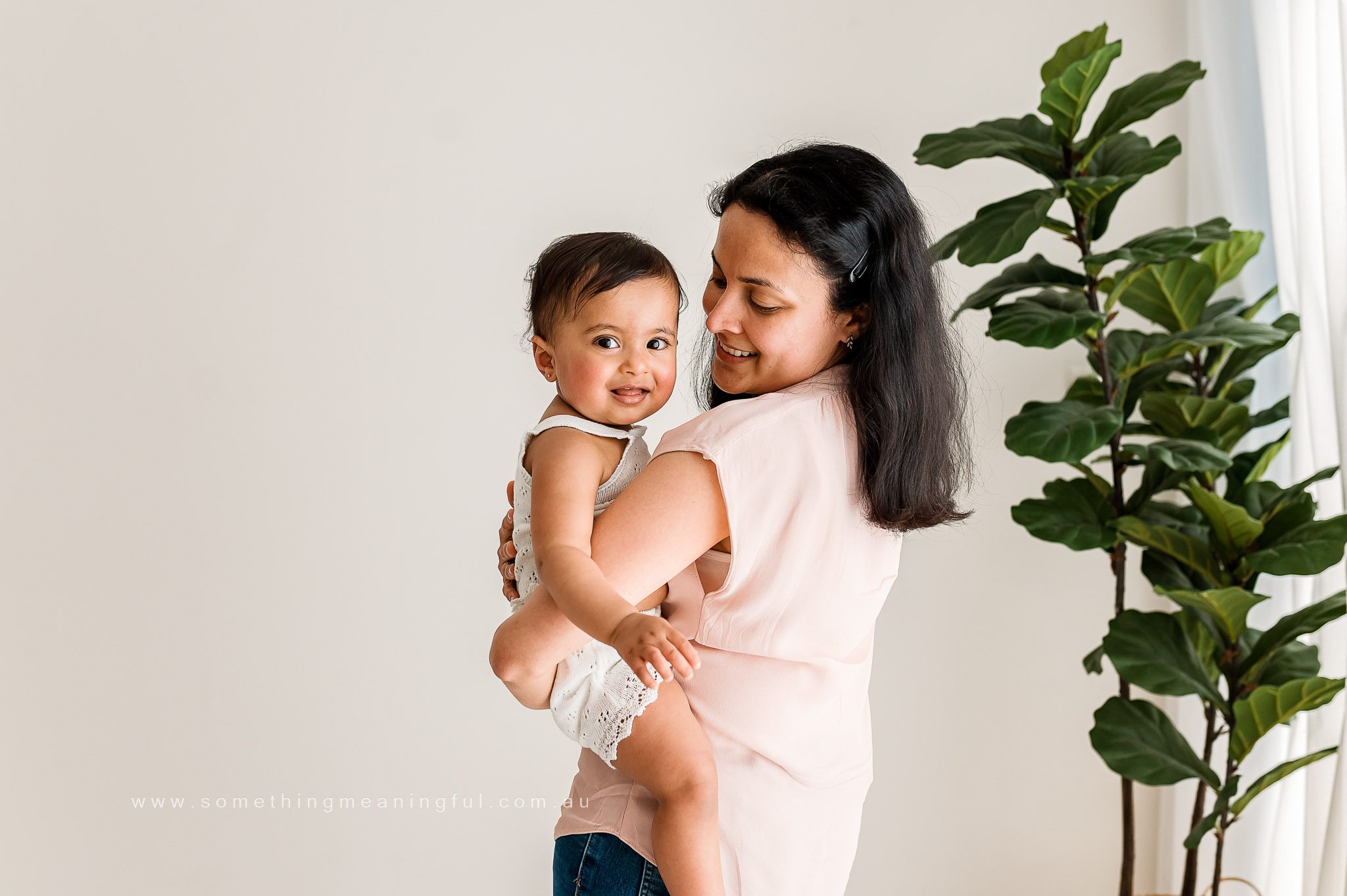 A woman smiles while holding a happy baby in her arms. Captured by a Melbourne family photographer, they stand indoors near a large green plant, with soft natural light coming through a window.