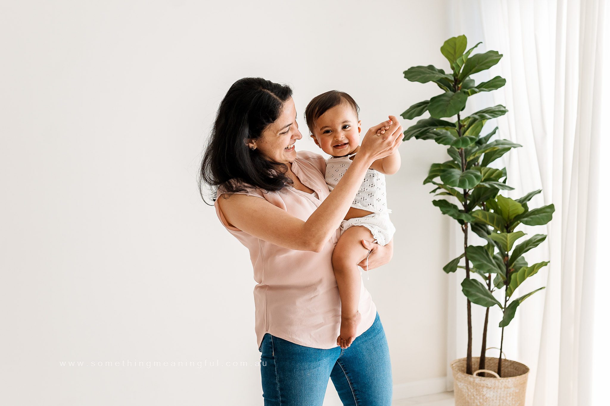A smiling woman in a sleeveless pink top holds a happy baby in her arms. Captured by a Melbourne family photographer, they stand indoors near a large green potted plant and window with sheer white curtains in a bright, minimalistic setting.