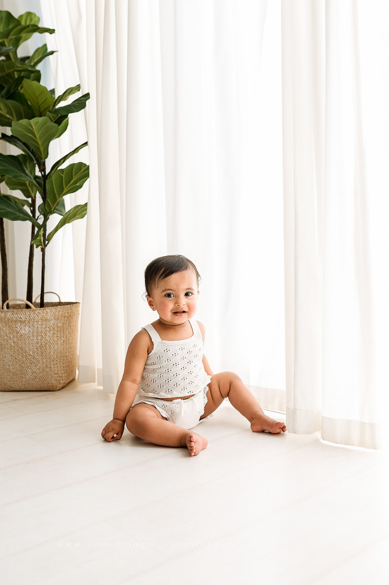 A smiling baby in a white outfit sits on a light-colored floor near sheer white curtains, with a large green plant in a woven basket in the background—captured by a Melbourne family photographer.