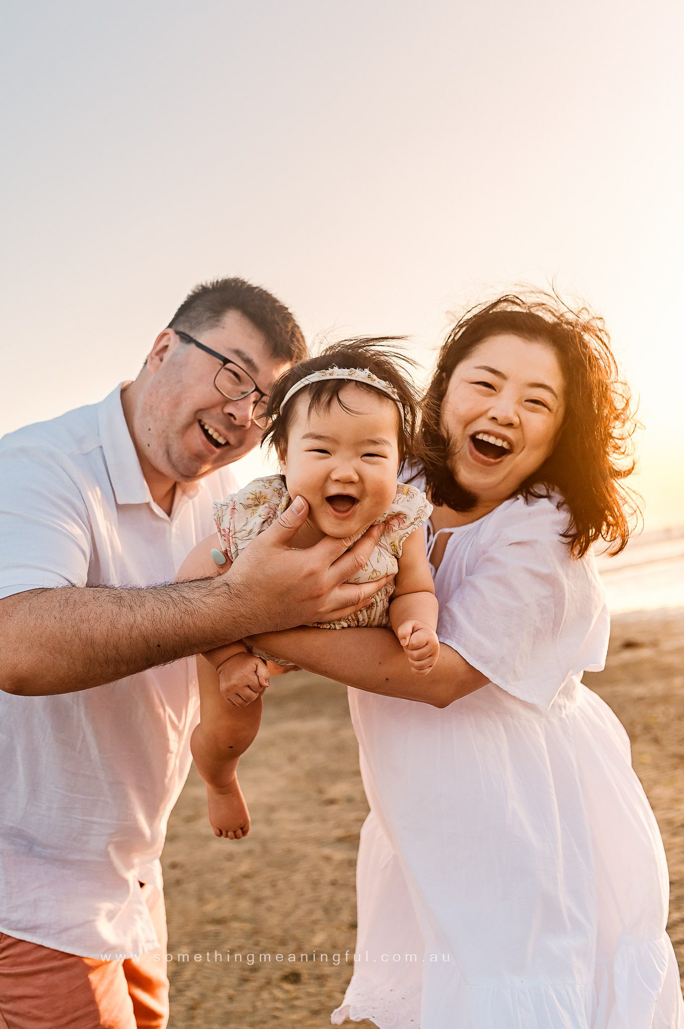 A joyful family of three, captured by a Family and Newborn Photographer Melbourne West, poses together on a sandy beach at sunset, with the man and woman holding their smiling baby and all dressed in light-colored clothing.