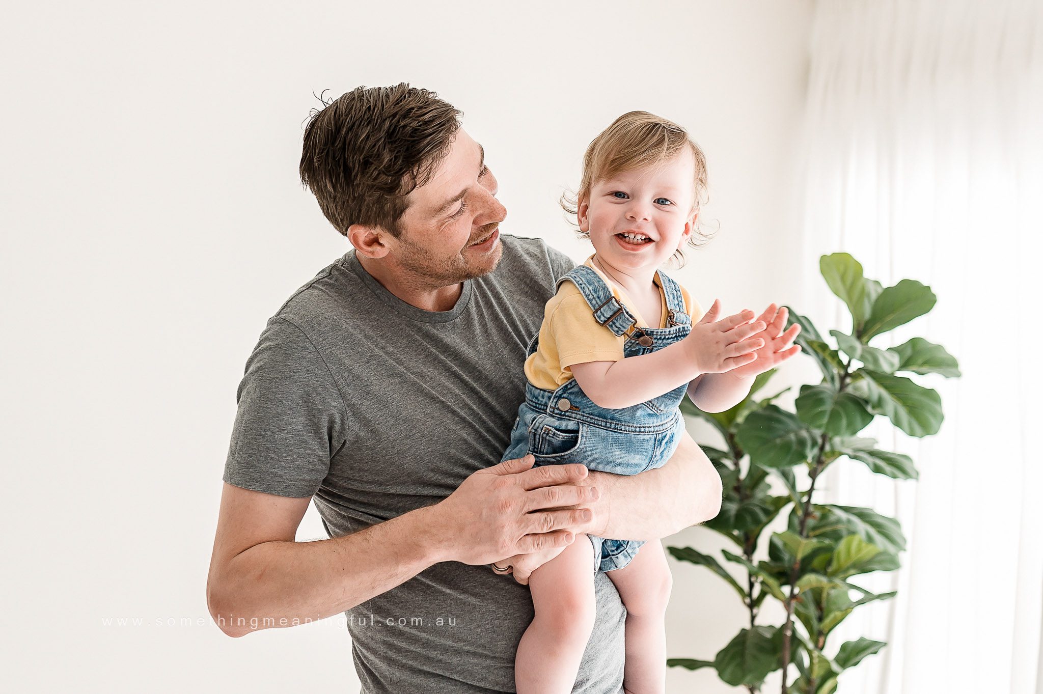 A smiling man in a grey t-shirt holds a laughing toddler in denim overalls and a yellow shirt. Captured by a Melbourne family photographer, they are indoors near a large green plant with light streaming through sheer curtains.