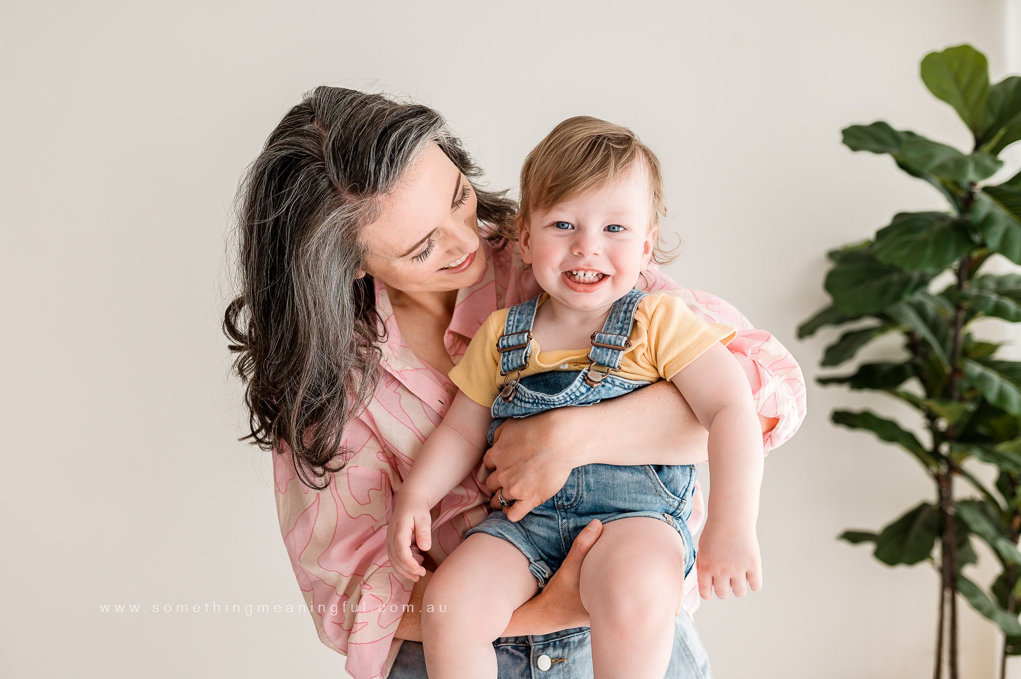 A smiling woman with gray-streaked hair in a pink blouse holds a cheerful toddler in denim overalls and a yellow shirt. Captured by a Melbourne family photographer, they pose indoors beside a leafy green plant.
