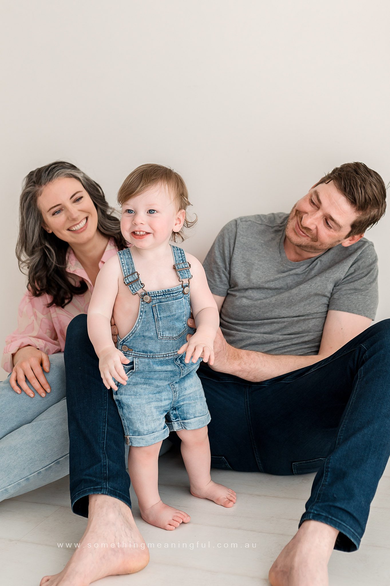 A smiling toddler in denim overalls stands barefoot between a seated woman with gray hair and a man in jeans, all posing against a light background—captured beautifully by a Melbourne family photographer.