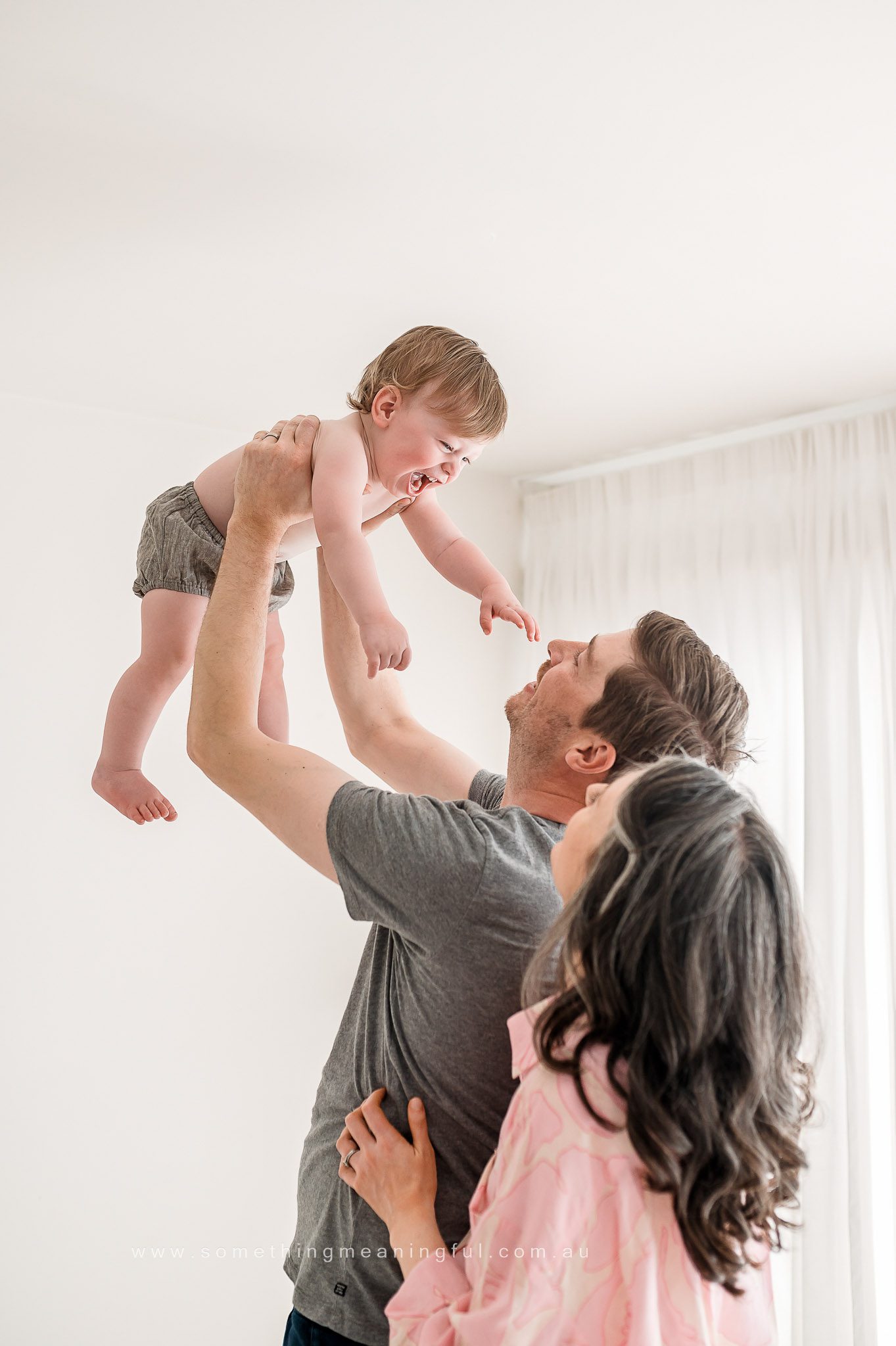 A man lifts a laughing baby into the air while a woman with long hair, standing beside him, smiles and gently touches his back in a bright, softly lit room—captured perfectly by a Melbourne family photographer.