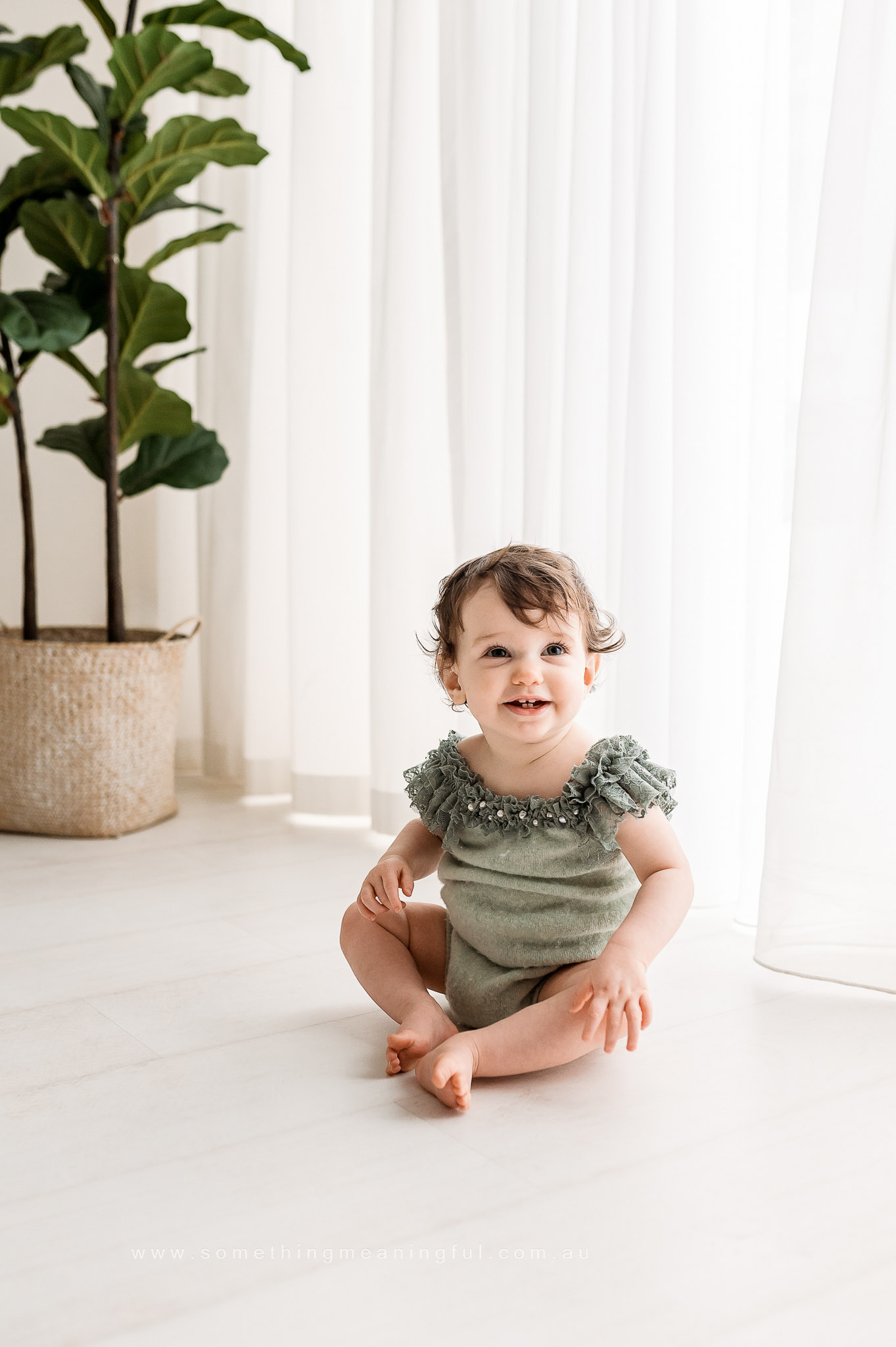 A smiling toddler with curly hair sits barefoot on a light floor, wearing a mint green ruffled outfit—perfect for family photography with baby. A tall potted plant and sheer white curtains brighten the airy room in the background.