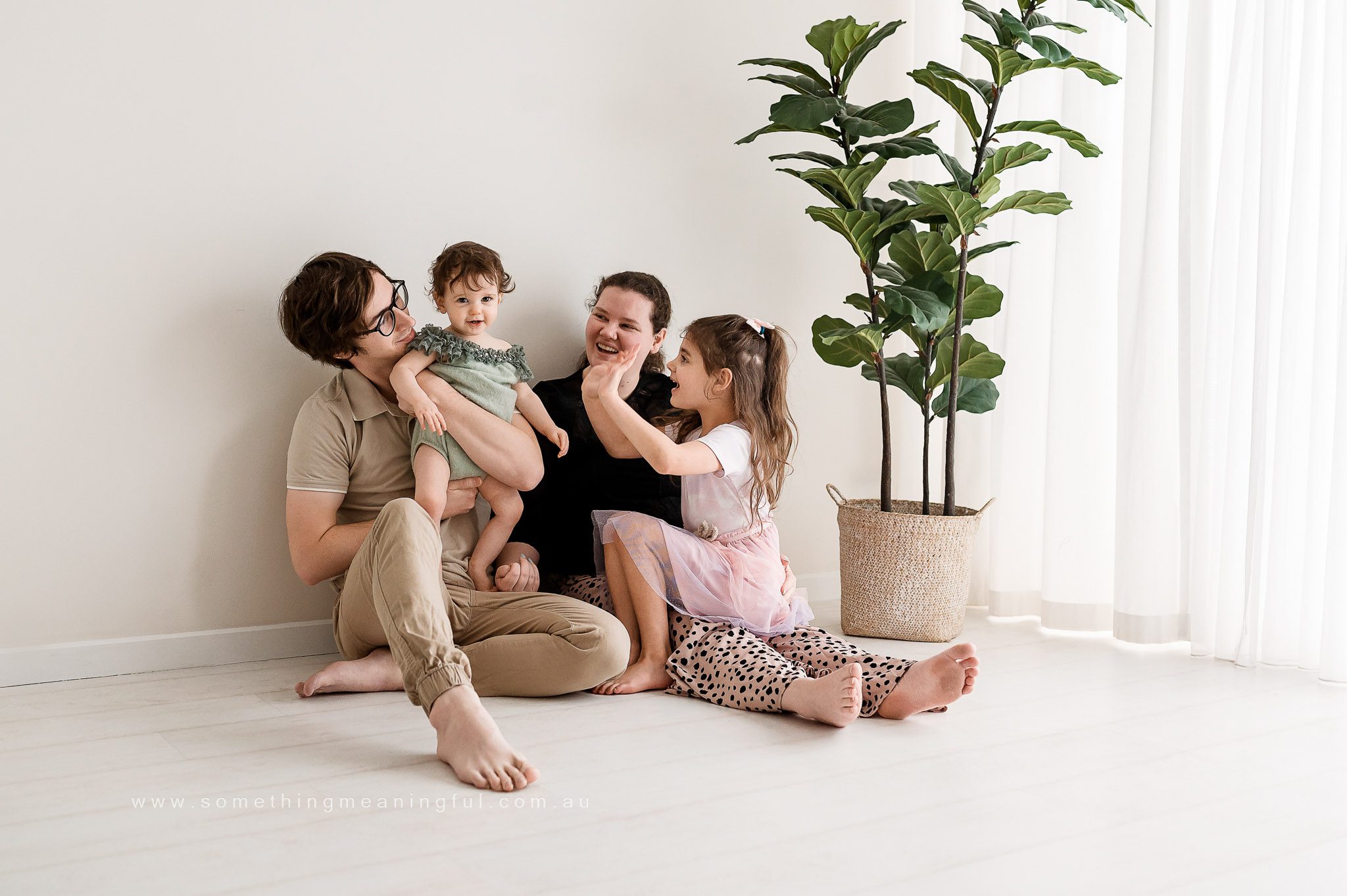 A family of four sits on the floor by a large potted plant in a bright, minimal room. The parents smile and interact with their two young children, capturing a cheerful, loving moment of family photography with baby.
