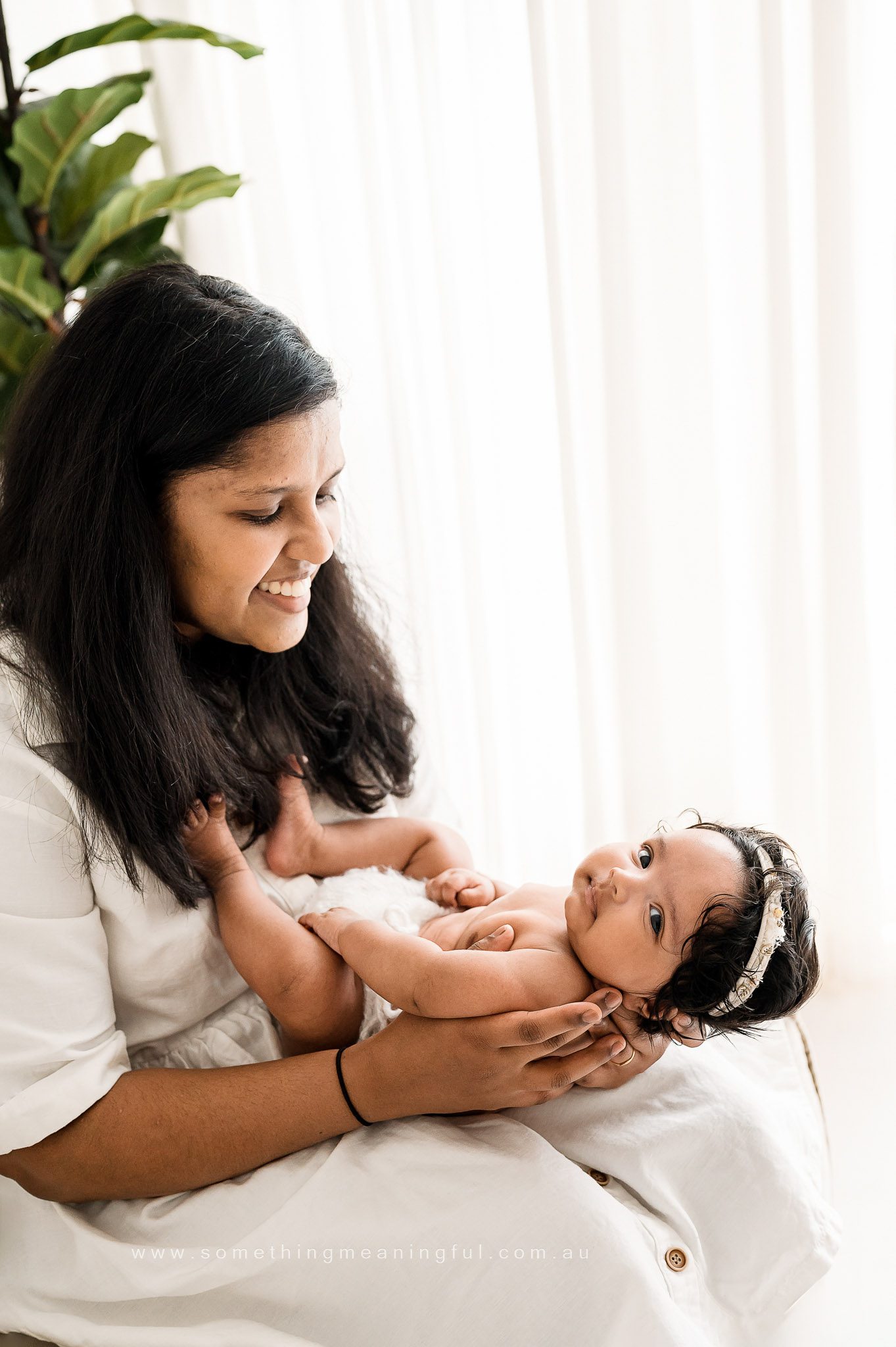 A woman with long dark hair smiles lovingly at a baby in her lap during a family photography with baby session. The baby, in a diaper and headband, gazes up calmly as soft natural light streams in through a nearby window.
