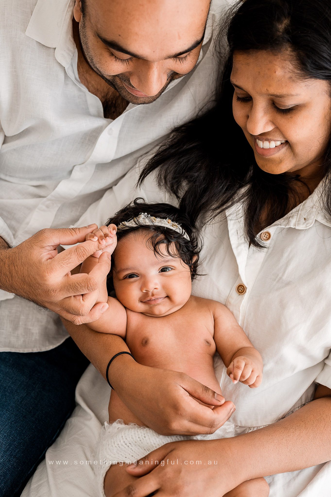 A smiling baby lies between two adults, likely parents, who are looking down at the baby lovingly. The baby reaches up while the adults gently hold the baby's hand—a heartwarming moment of family photography with baby, all dressed in light-colored clothing.