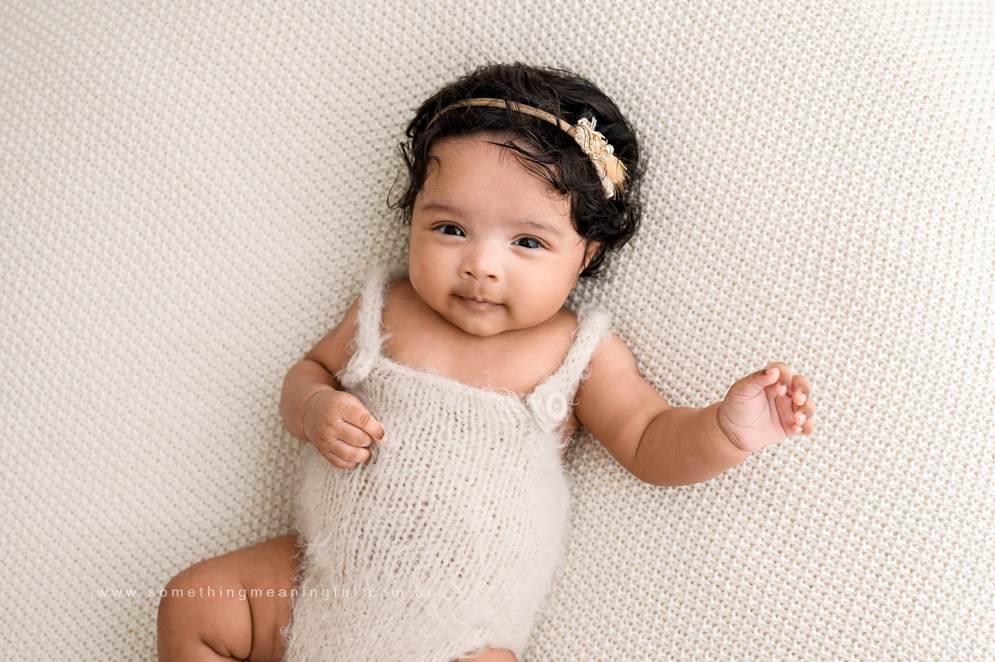 A baby with dark hair and a gold headband lies on a cream-colored knitted blanket, wearing a soft, white fuzzy outfit, and looks at the camera with a gentle smile—perfect for family photography with baby.