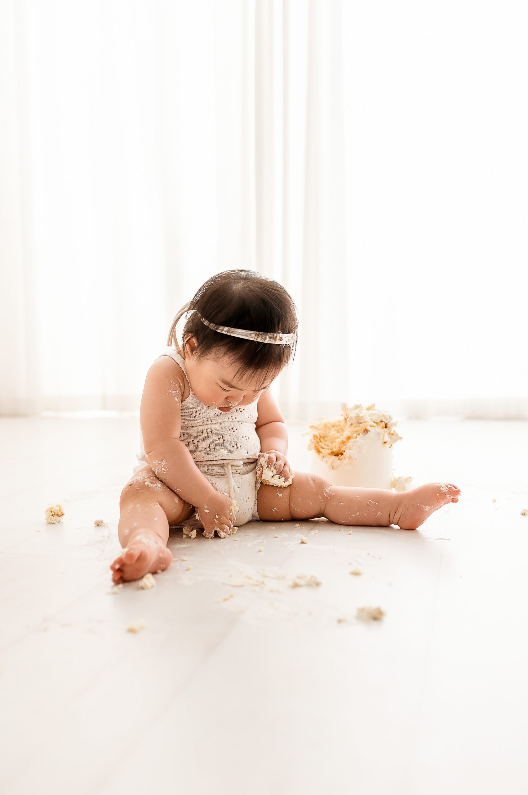 A baby wearing a white outfit and a headband sits on the floor, playing with pieces of cake. There are cake crumbs scattered around and a partially eaten cake on the floor nearby. The background is bright and softly lit. Captured by Family and Newborn Photographer Melbourne West 