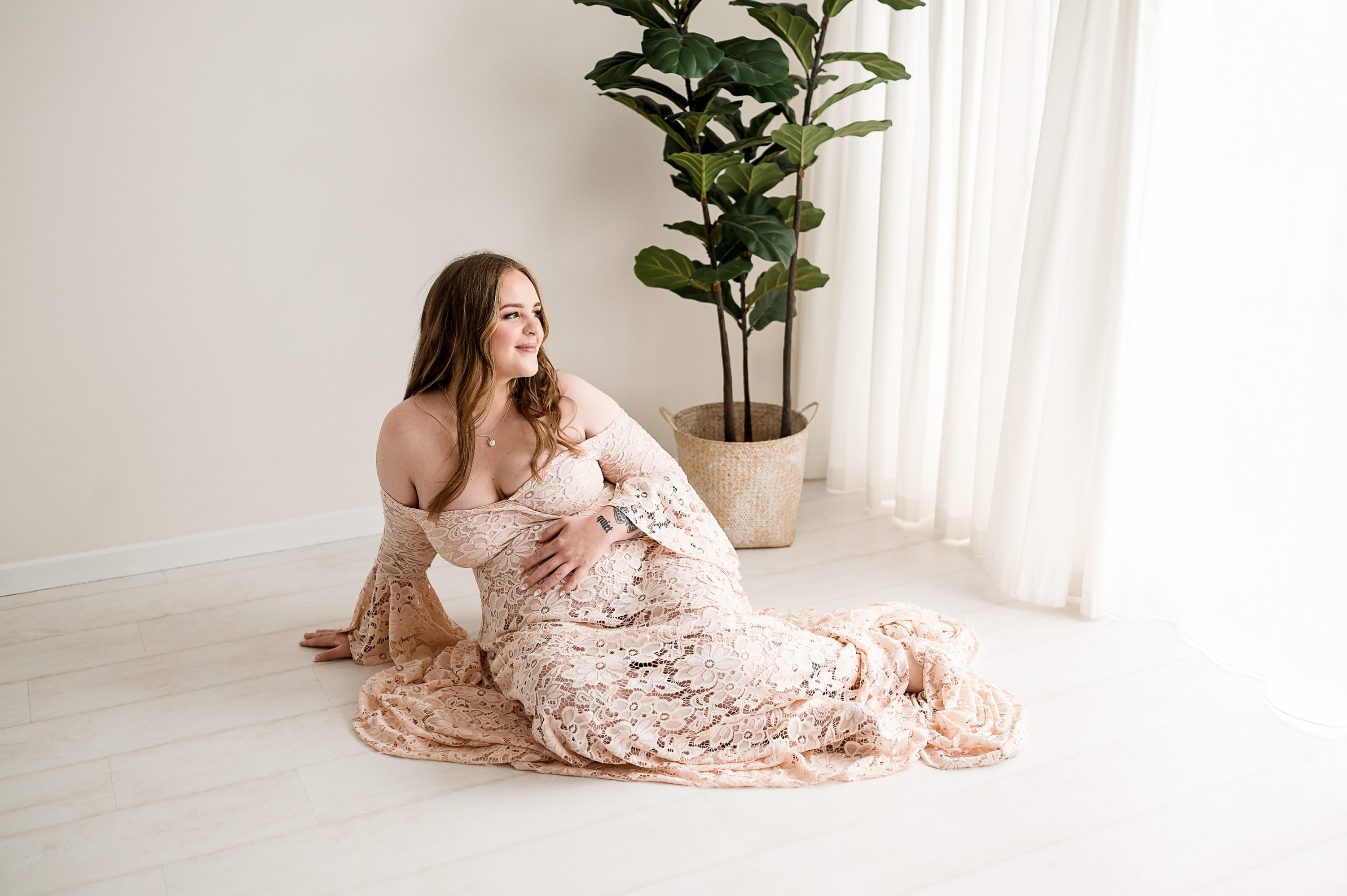 A pregnant woman in a lacy, off-the-shoulder gown sits on a light floor, holding her belly during Maternity Photography Melbourne West. She smiles while looking out a sunlit window during her maternity studio photoshoot, with a tall potted plant in the background.