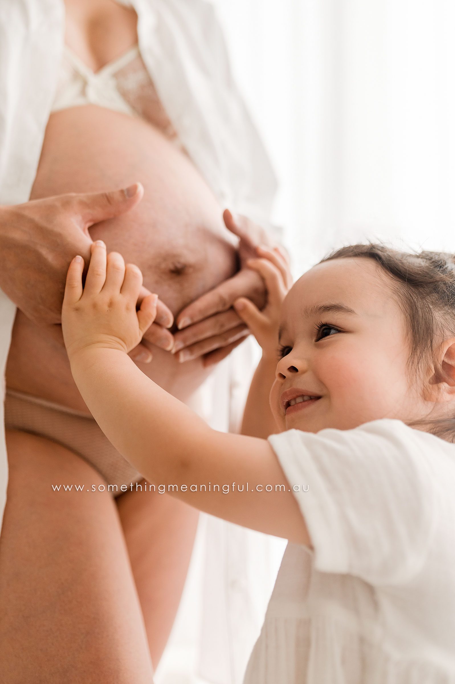A smiling young child gently touches the bare pregnant belly of their mother, during a tender and joyful maternity studio photoshoot. Both wear light clothing in a bright, airy room.