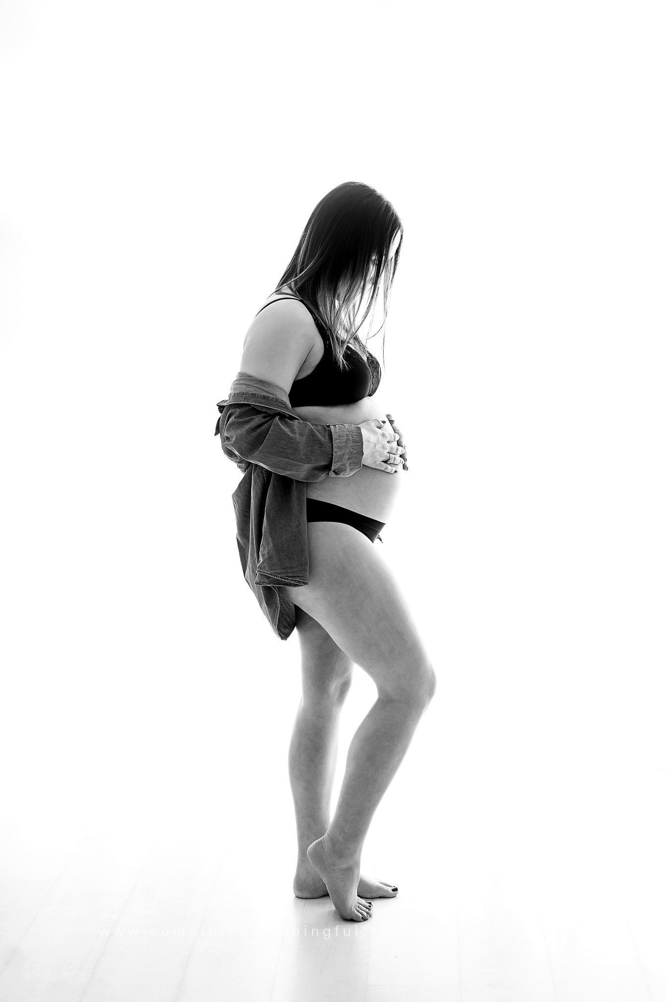 A pregnant woman in underwear and an open shirt stands barefoot, looking down and gently holding her belly during a bright white maternity studio photoshoot.