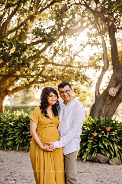 A smiling couple stands outdoors in sunlight, surrounded by greenery—one of the most charming photography session locations. The woman in a mustard yellow dress is visibly pregnant as the man in glasses gently holds her baby bump.