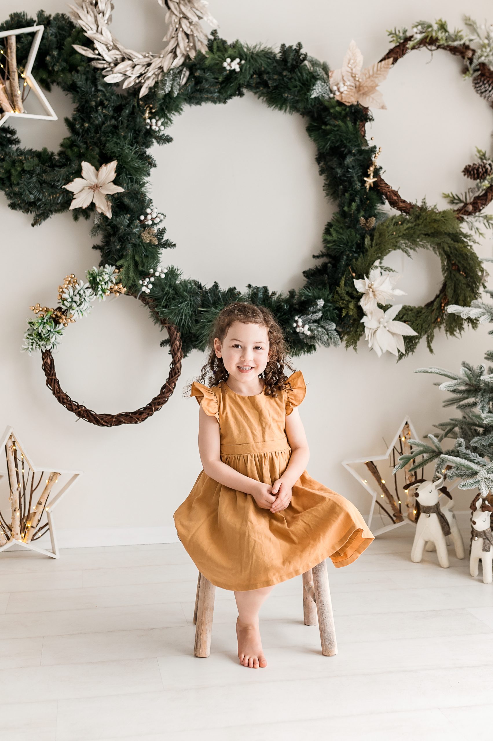 A young girl in a mustard yellow dress sits barefoot on a wooden stool, smiling. Behind her, a white wall is adorned for a Christmas mini session in Melbourne’s west, with green wreaths, white poinsettias, stars, and festive decorations.