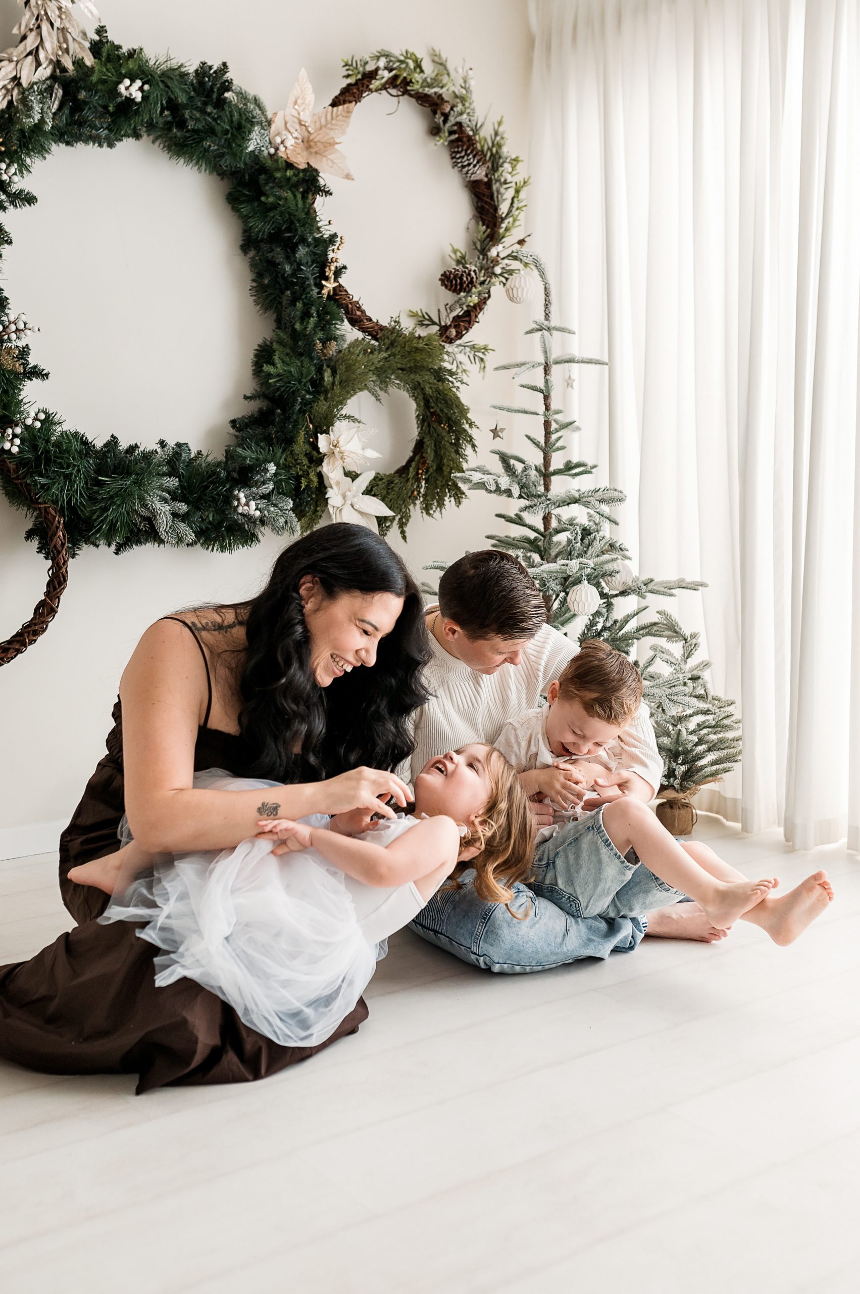 A joyful family of four, including two young children, sits on the floor in a bright room with large wreaths and a small Christmas tree. They are smiling and playing together during a festive Christmas mini session in Melbourne’s west.
