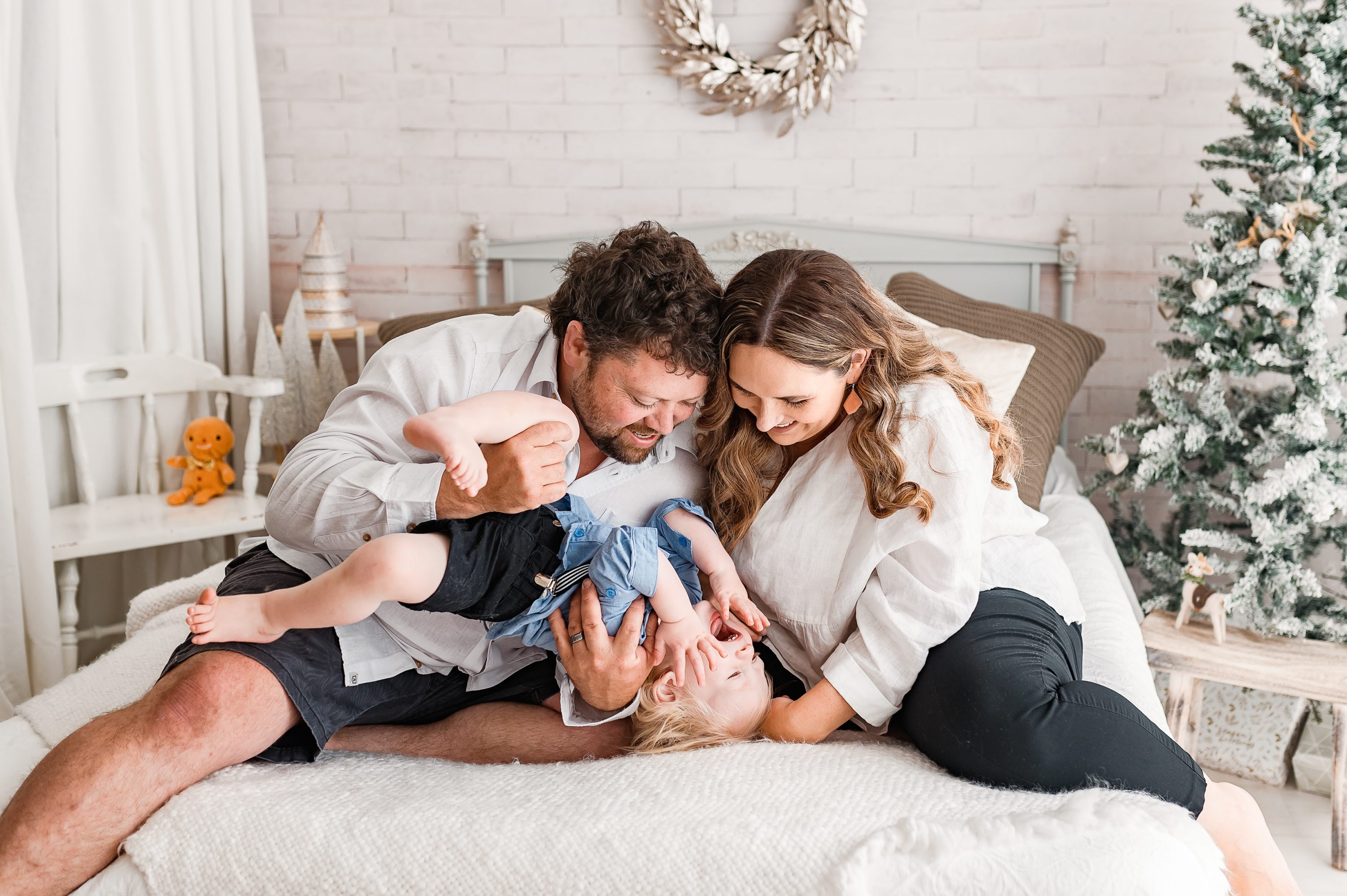 A smiling couple sits on a bed in a cozy, light-filled room, playfully tickling a young child who is lying upside down. A decorated Christmas tree and holiday decorations set the scene for a joyful Christmas mini session in Melbourne’s west.