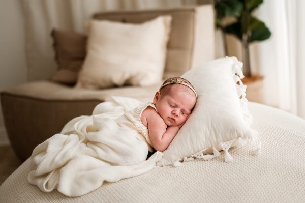 A Family and Newborn Photographer Melbourne West captures a peaceful baby wearing a headband, sleeping soundly on a white pillow, wrapped in a soft cream blanket, with a cozy chair and pillow softly blurred in the background.