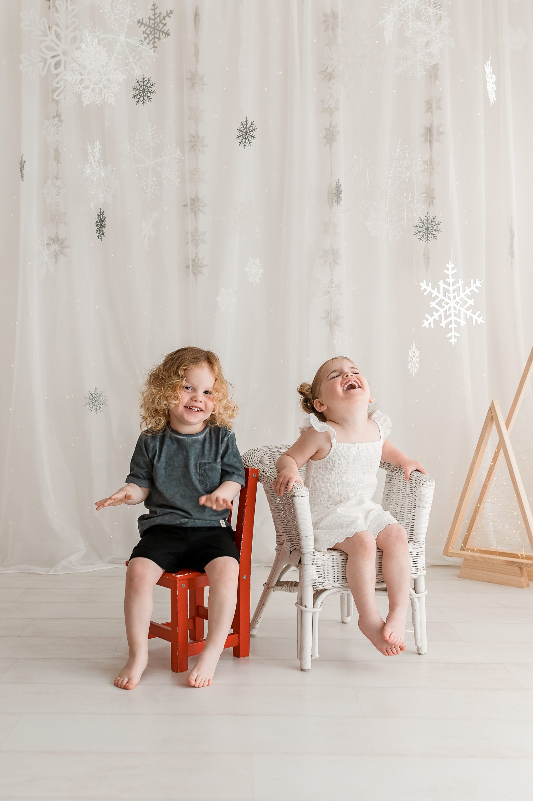 Two young children sit barefoot on chairs in a bright room with snowflake decorations hanging behind them, sharing a joyful moment during a Christmas mini session in Melbourne’s west.