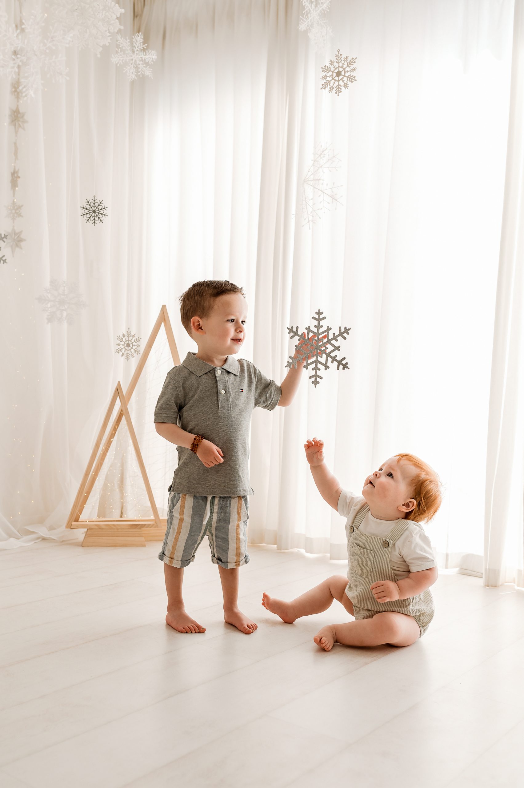 Two young children, one standing and one sitting, play with hanging decorative snowflakes in a bright room—perfect for a Christmas mini session in Melbourne’s west. White curtains and a wooden triangular decor piece brighten the backdrop.