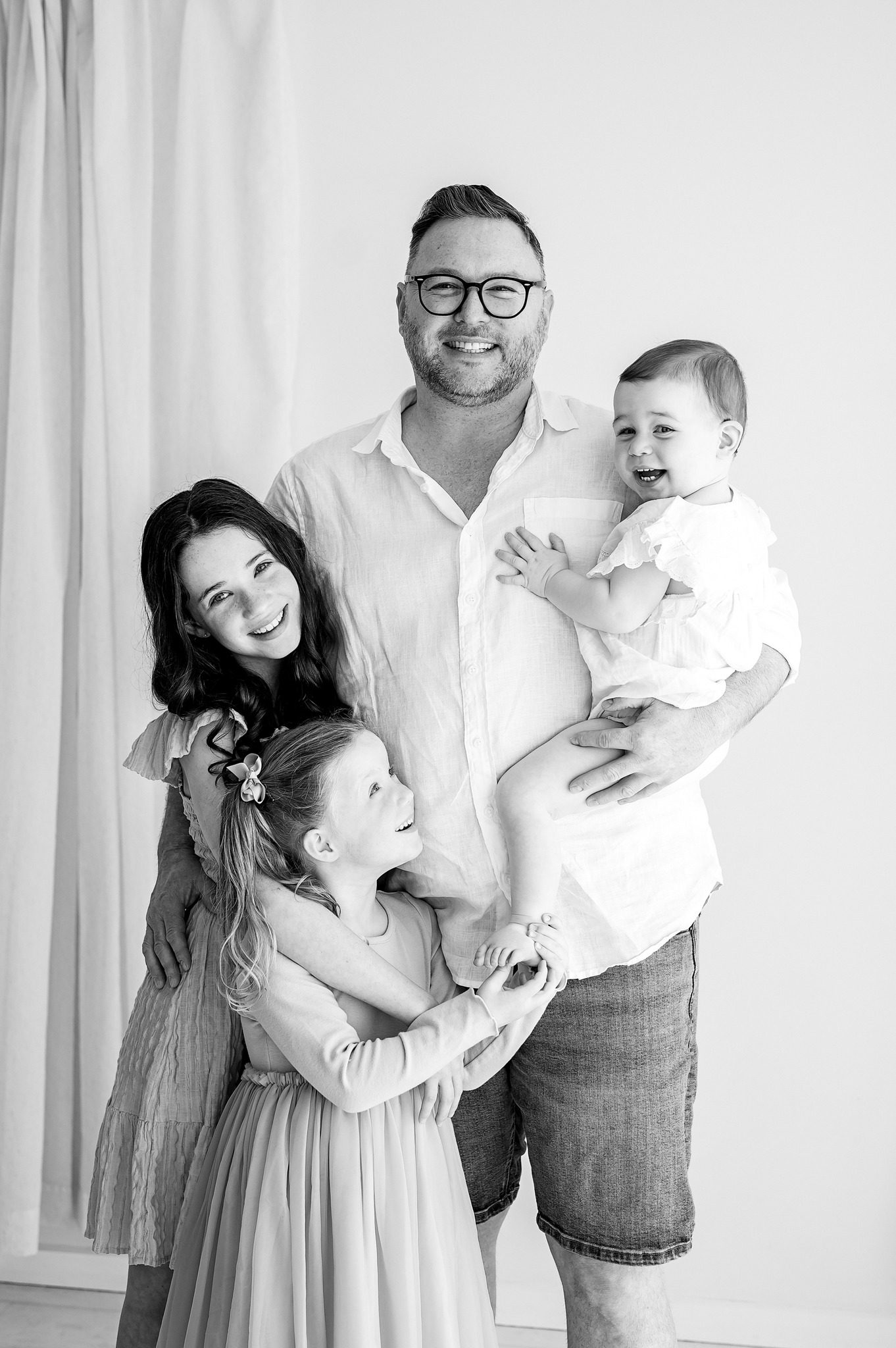 A smiling man in glasses holds a baby while standing with two young girls during a family mini session Melbourne. Everyone is dressed in light-colored clothes and looks happy. The photo is in black and white with a simple, bright background.