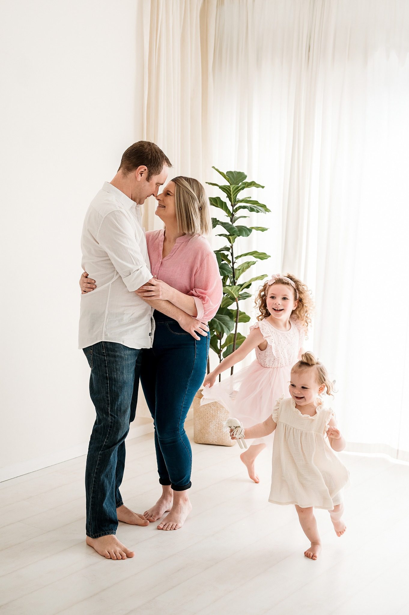 A couple embraces and smiles at each other in a bright room during their family mini session Melbourne, while two young girls in light dresses joyfully run and play around them. A green potted plant sits by a window with sheer curtains.