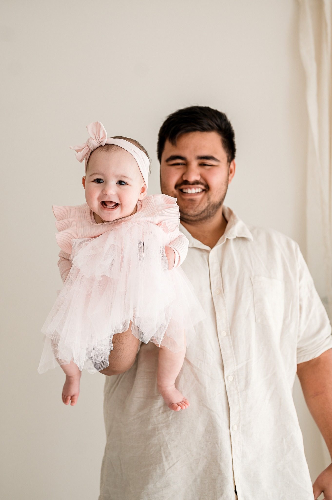 A man in a light-colored shirt smiles while holding a happy baby girl in a pink dress and headband. The plain, light background creates a soft, cheerful scene—perfect for capturing memories at a family mini session Melbourne.