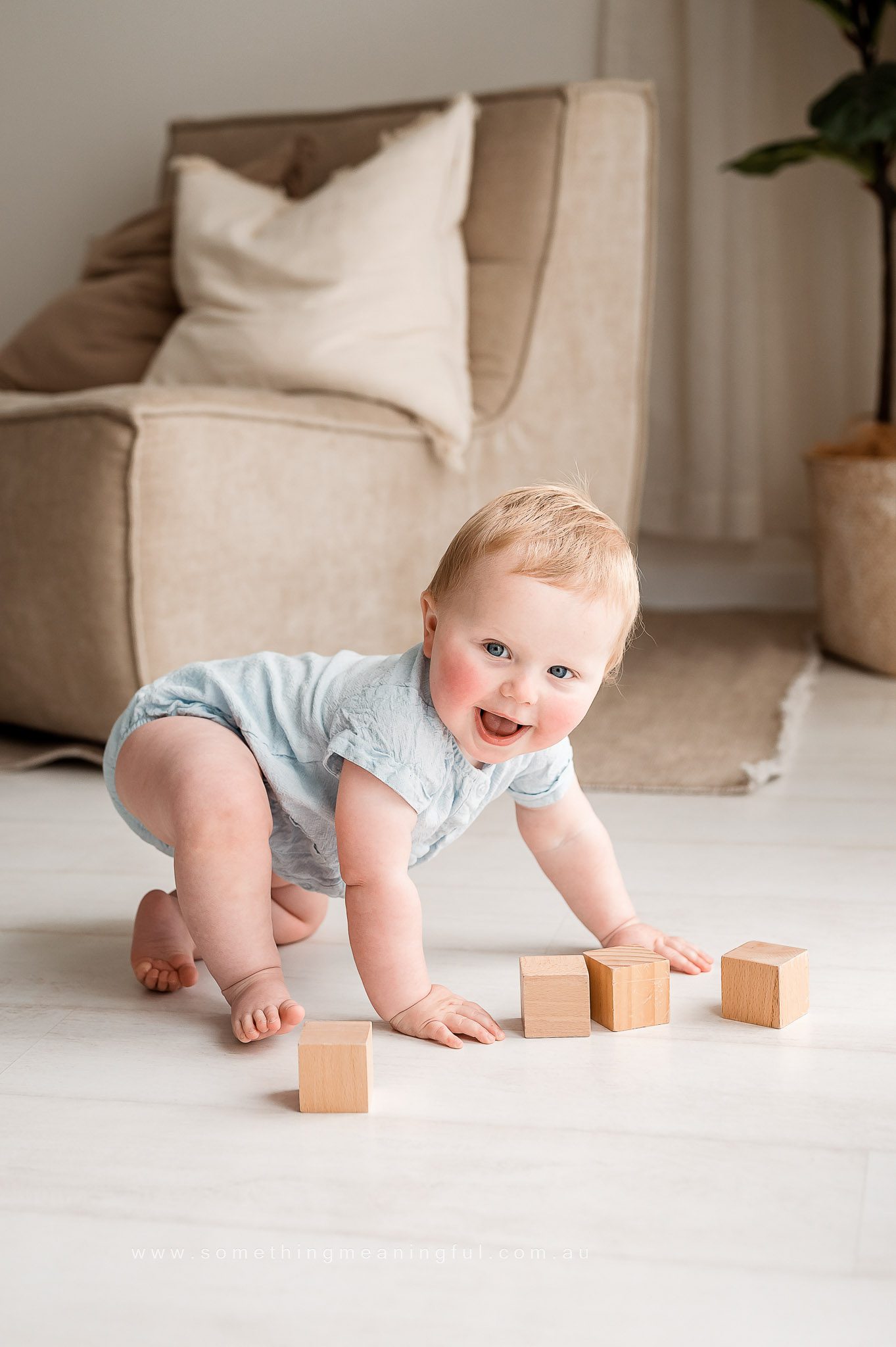 A smiling baby in a light blue outfit crawls on a white floor, playing with four wooden blocks—perfect for capturing precious moments at a family mini session Melbourne. A beige armchair and a plant in a basket are softly visible in the background.