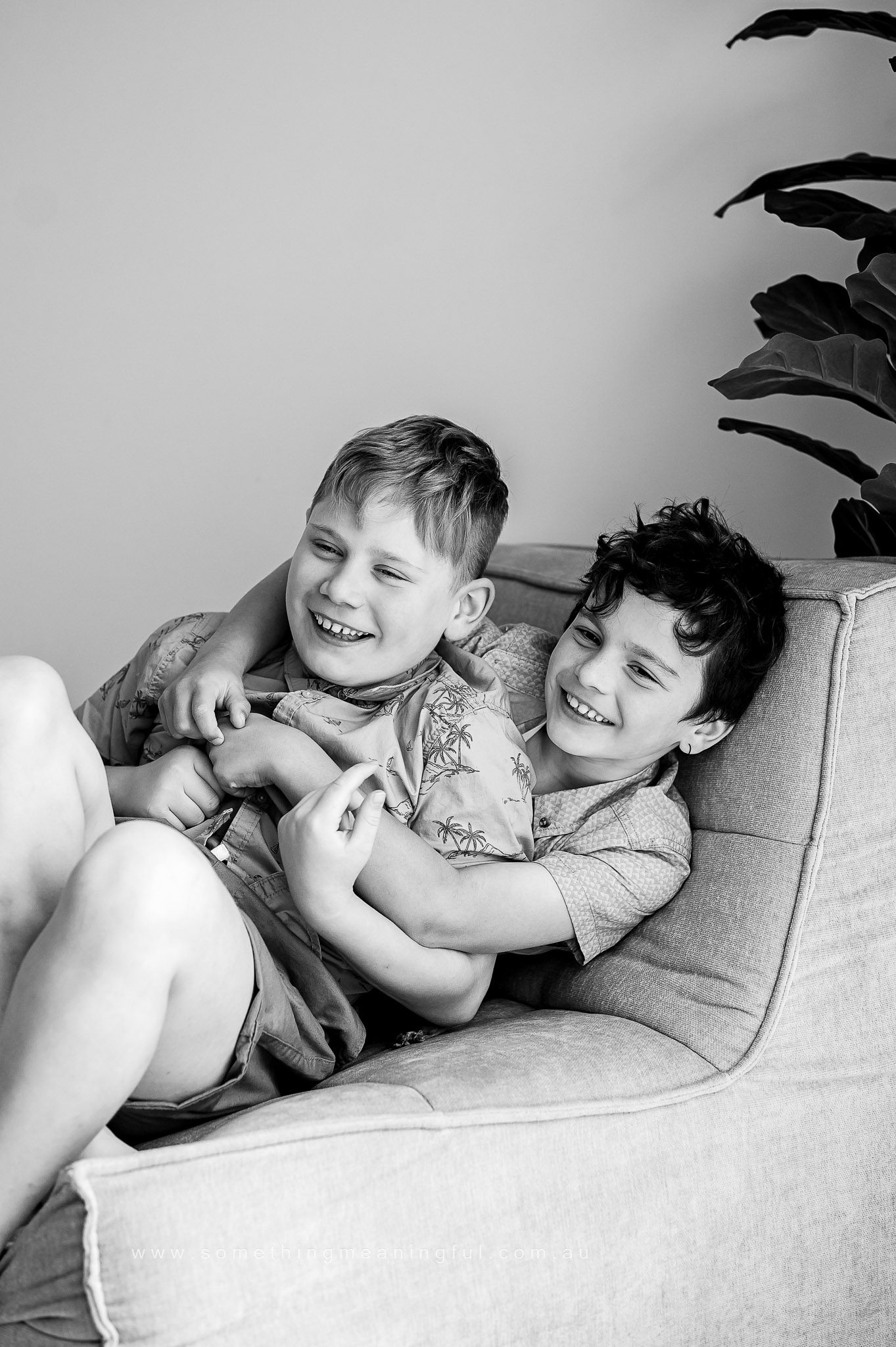 Two young boys sit on a couch, laughing and hugging each other playfully during a family mini session in Melbourne. The black and white image features a large plant in the background.