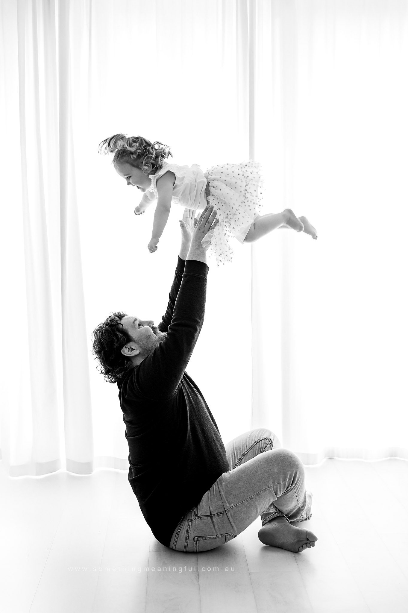 A man kneels on the floor and lifts a smiling young girl in a dress above his head, both enjoying a playful moment in a bright room with sheer curtains during their family mini session Melbourne.