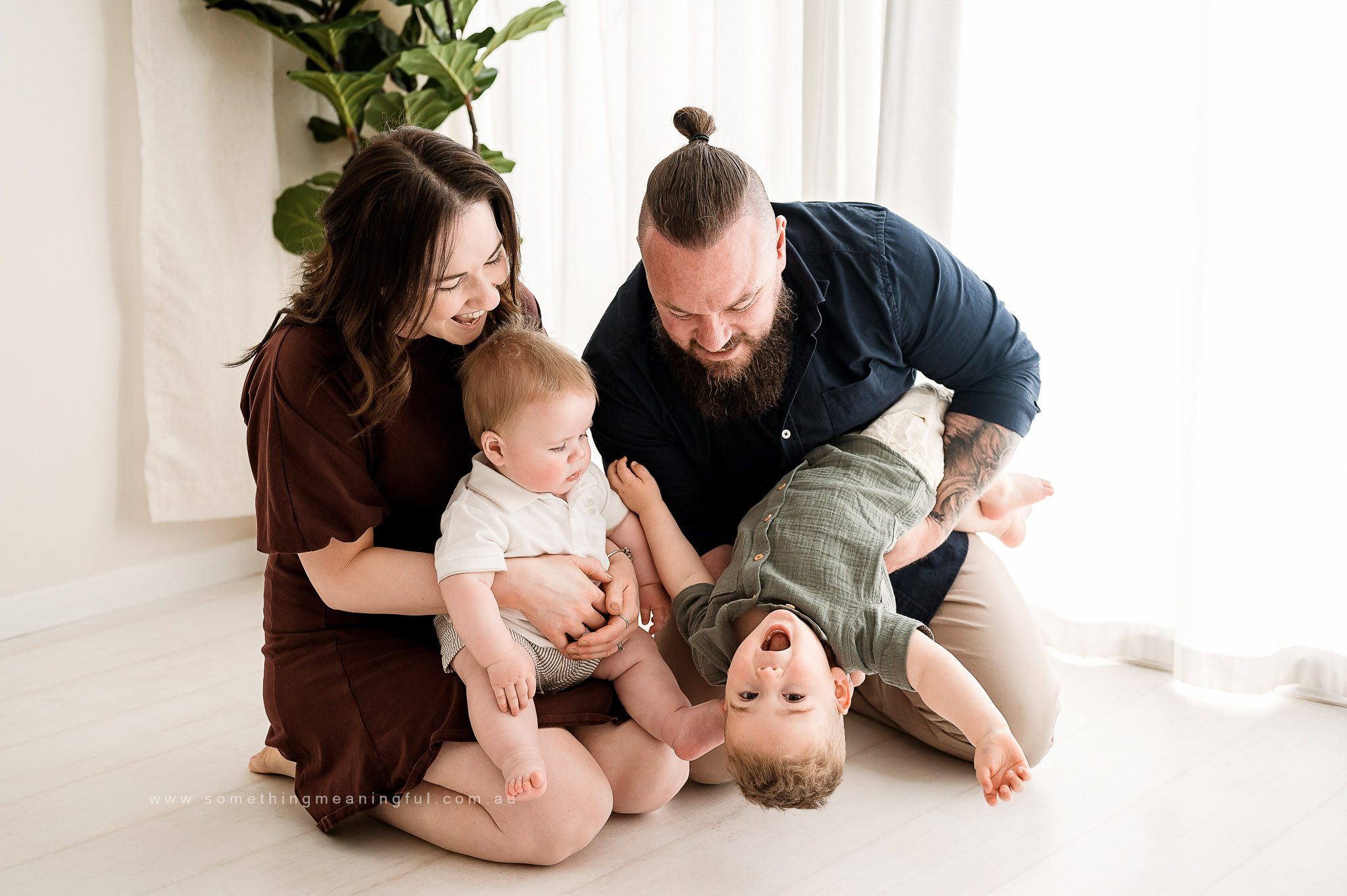 A smiling family of four sits on the floor, enjoying playful moments during our Fathers Day mini sessions. The mother holds a baby while the father holds a laughing toddler upside down, with bright natural light and a green plant in the background.