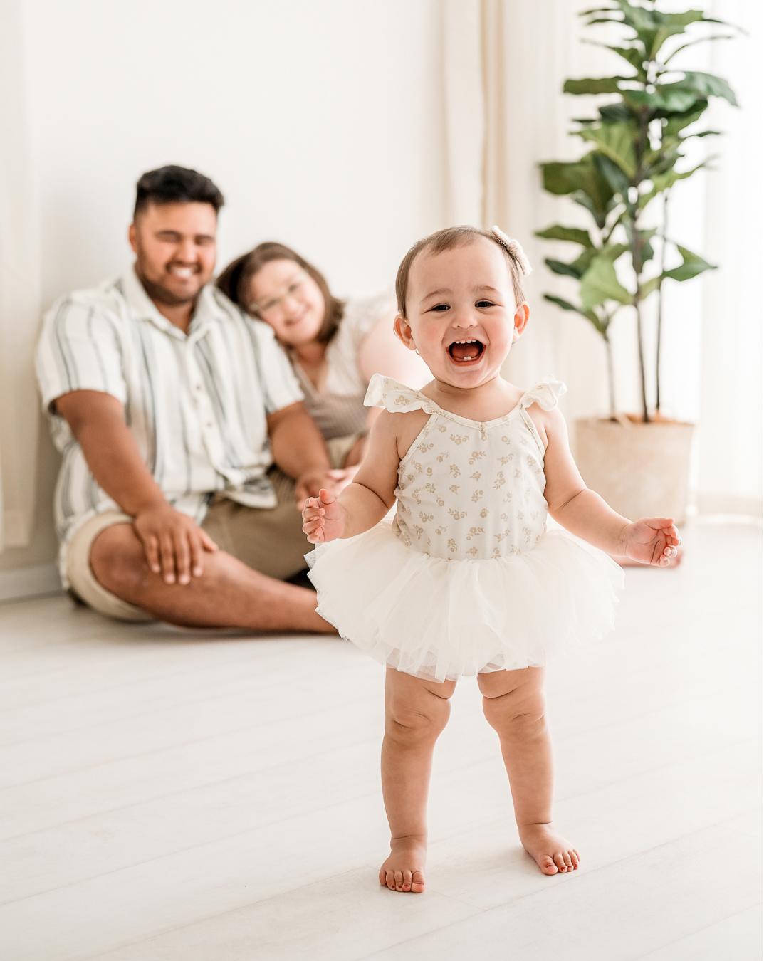 A smiling baby in a white dress stands in the foreground during a family mini session Melbourne, while two adults sit on the floor in the background, smiling beside a potted plant in a bright, airy room.