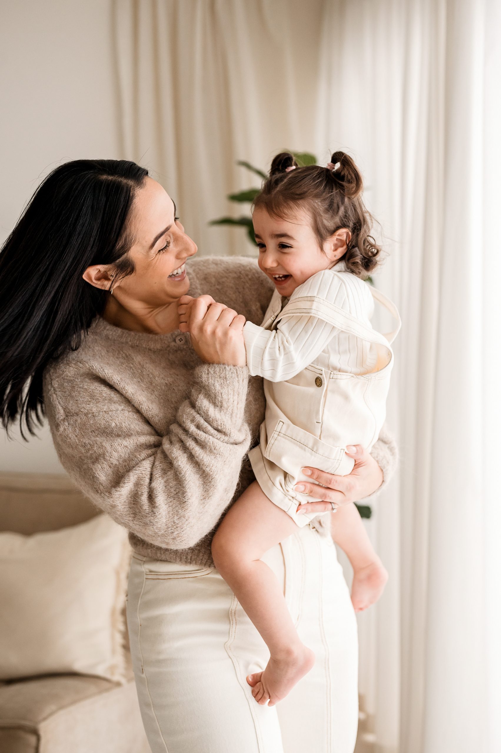 A woman with dark hair holds a smiling toddler in her arms indoors during a joyful mother's day mini session. Both wear light-colored clothing, sharing a happy moment together with light curtains and a plant in the background.