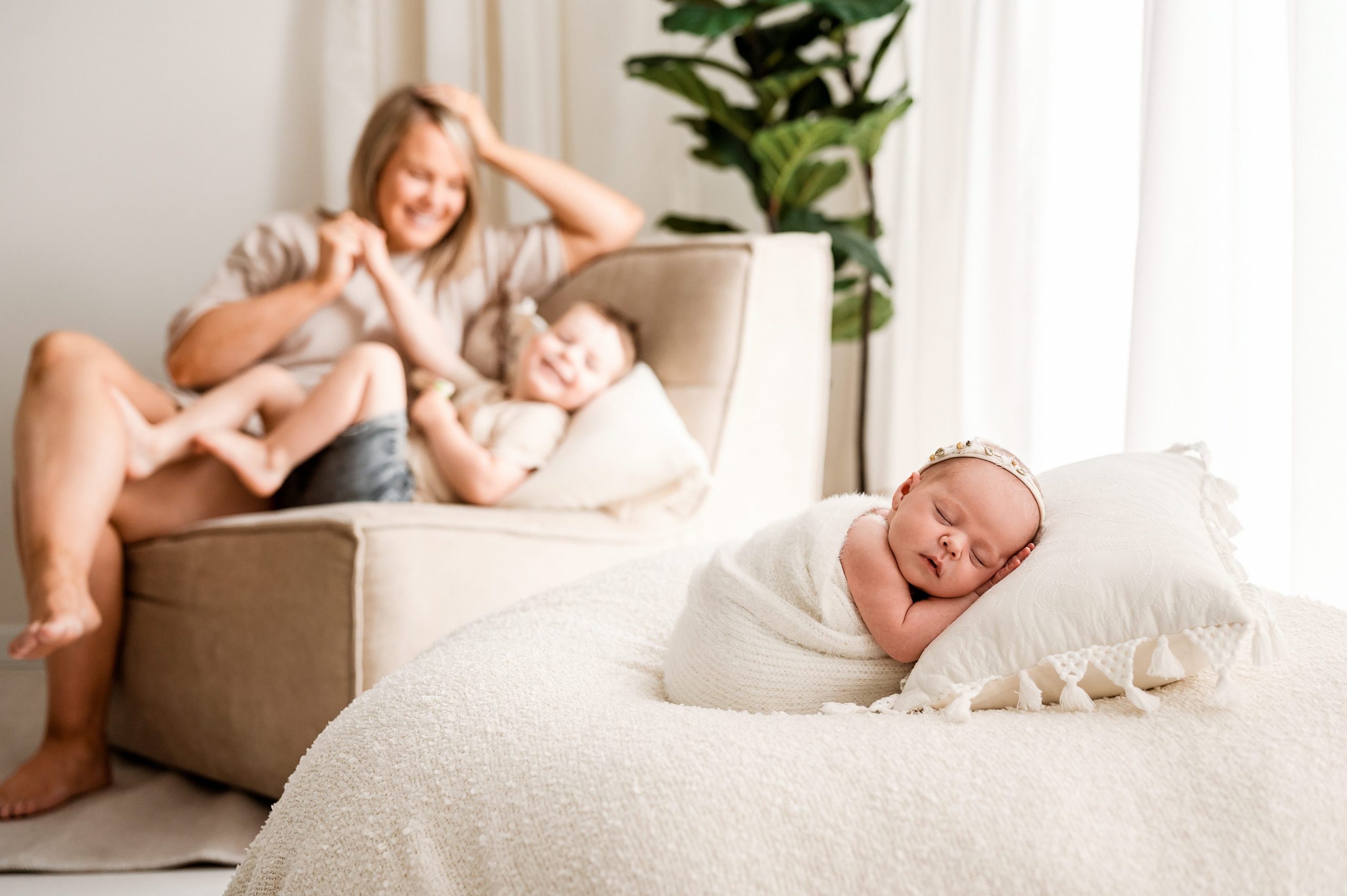 A newborn and family photographer Melbourne captures a baby wrapped in a white blanket sleeping peacefully, while a smiling woman and young child play together on a beige chair in the softly lit background.