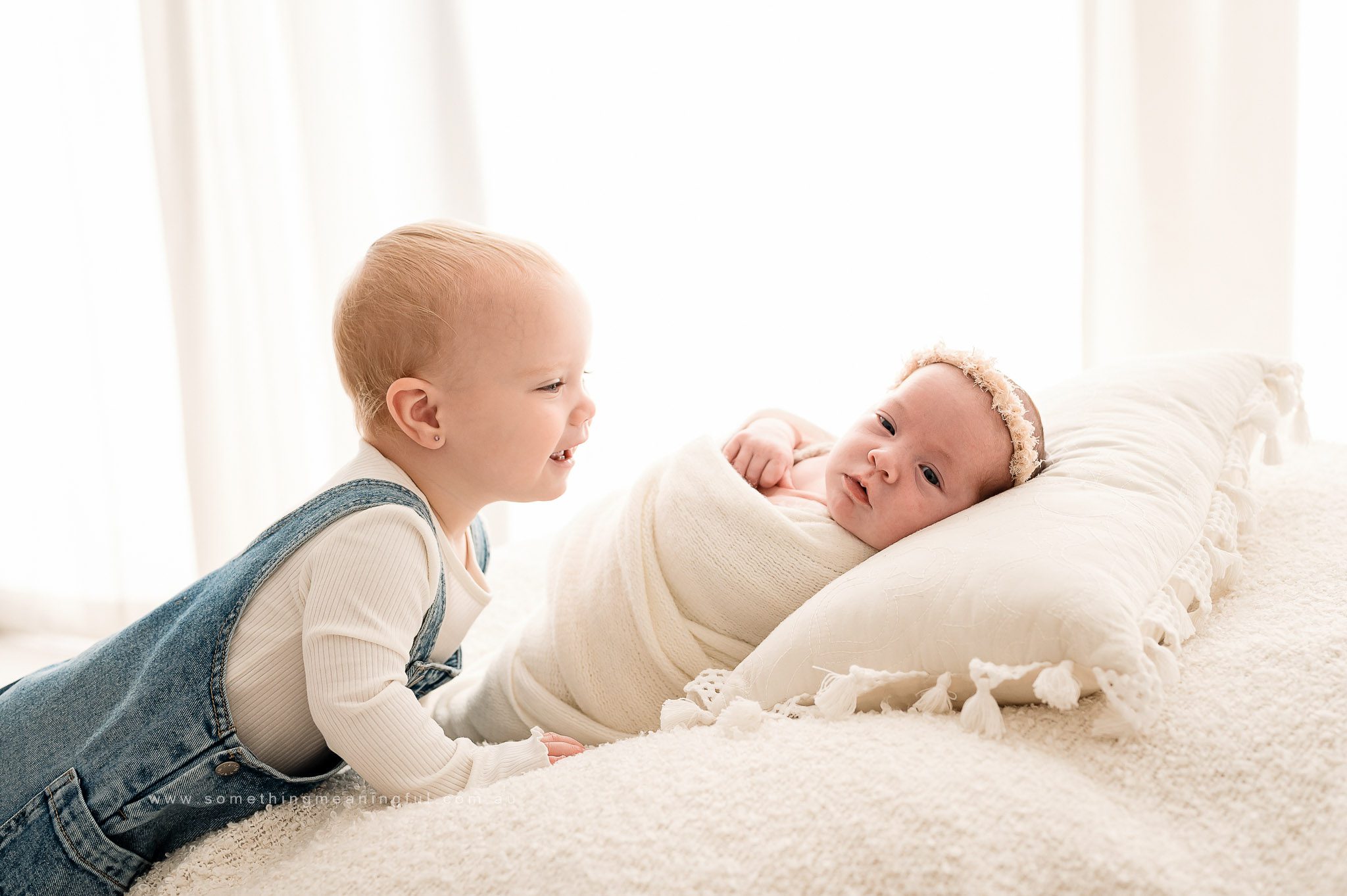 A toddler in denim overalls smiles at a newborn baby wrapped in a white blanket on a soft, white bed. Capturing precious Irish Twins Photography Melbourne moments, bright natural light fills the background.