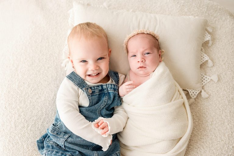 A smiling toddler in denim overalls sits next to a swaddled baby on a soft cream-colored blanket. Capturing Irish Twins Photography Melbourne style, the toddler holds the baby’s hand while the newborn wears a sweet headband.