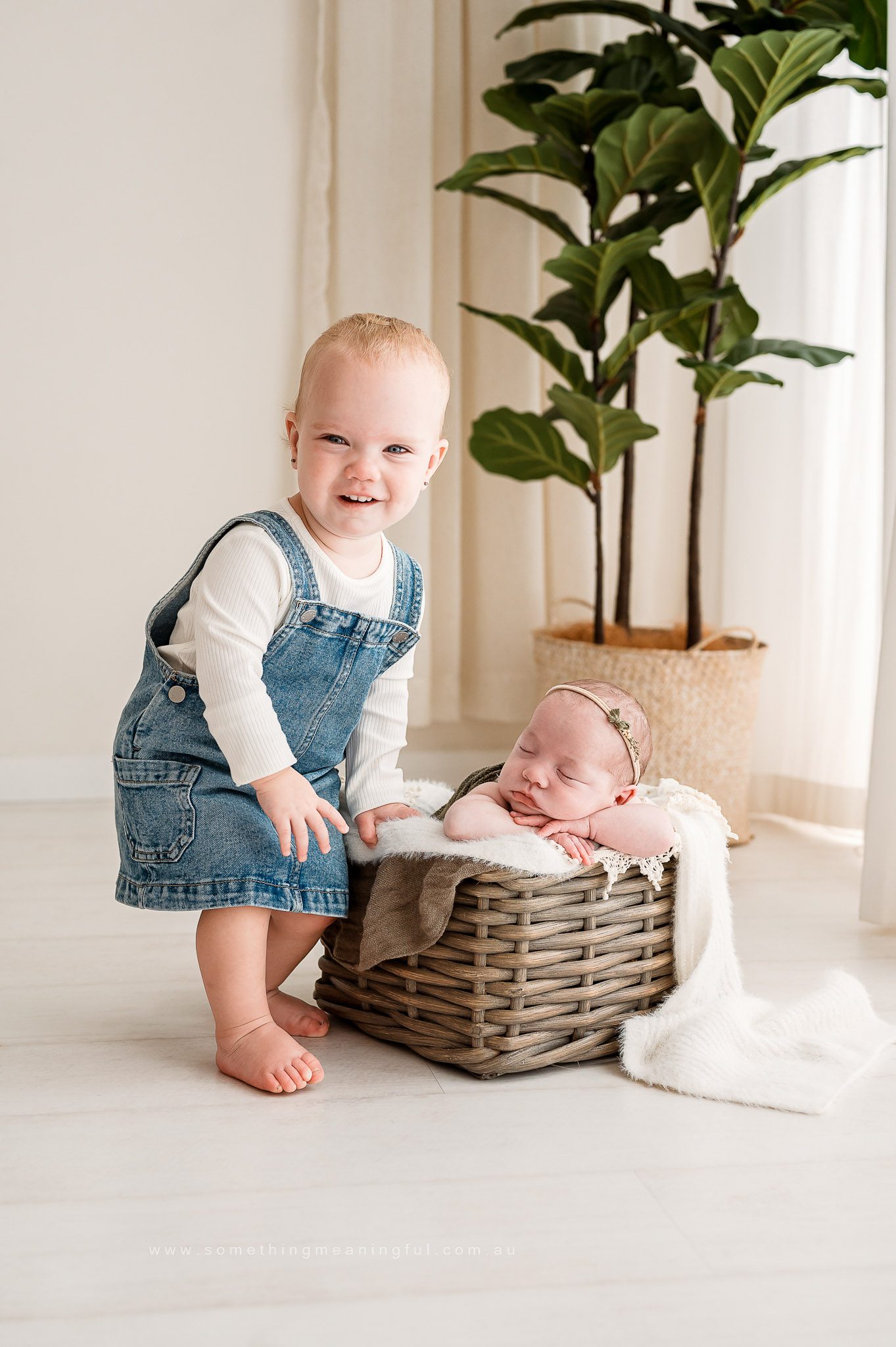A smiling toddler in a denim dress stands barefoot beside a wicker basket, where a newborn baby with a headband sleeps on a cozy blanket—an adorable Irish Twins Photography Melbourne session in a softly lit room with a tall green plant in the background. Captured by Family and Newborn Photographer Melbourne West