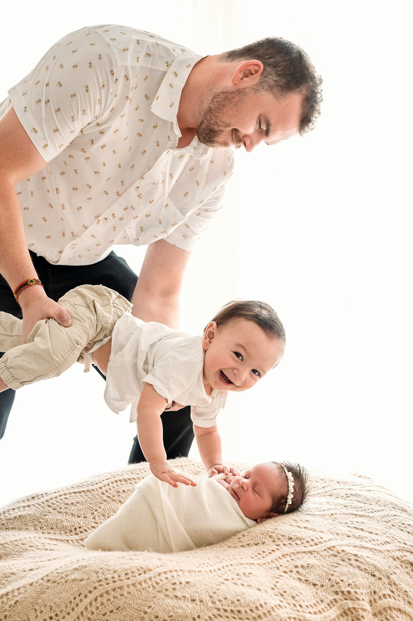 A smiling man holds up a laughing toddler, who reaches out to touch a newborn baby wrapped in a blanket and lying on a knitted coverlet. The scene is bright and joyful—perfect for Irish Twins Photography Melbourne.