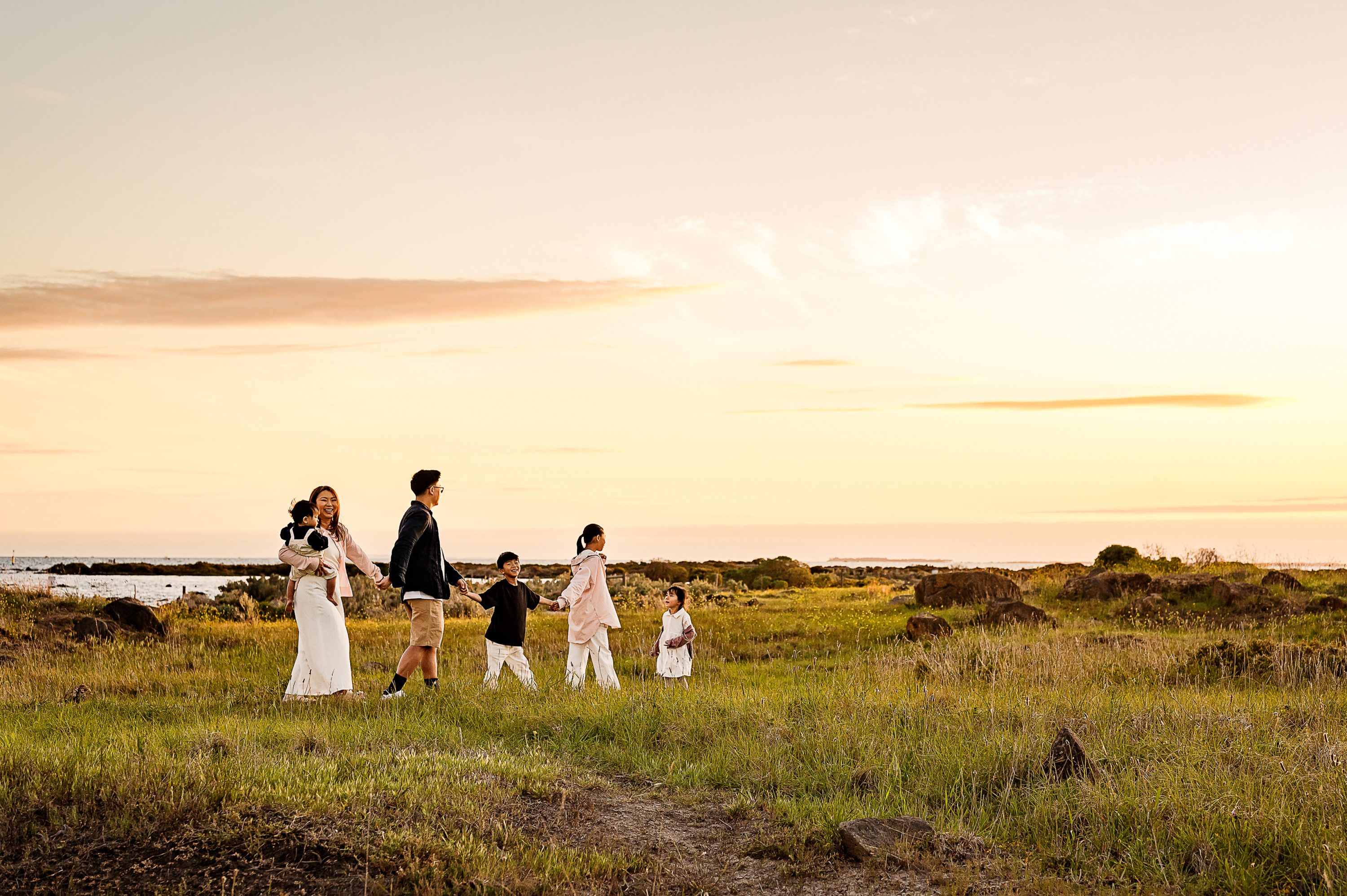 A family of six walks hand in hand through a grassy field at sunset, with the sky glowing and the ocean visible in the background—one of the most stunning photography session locations.