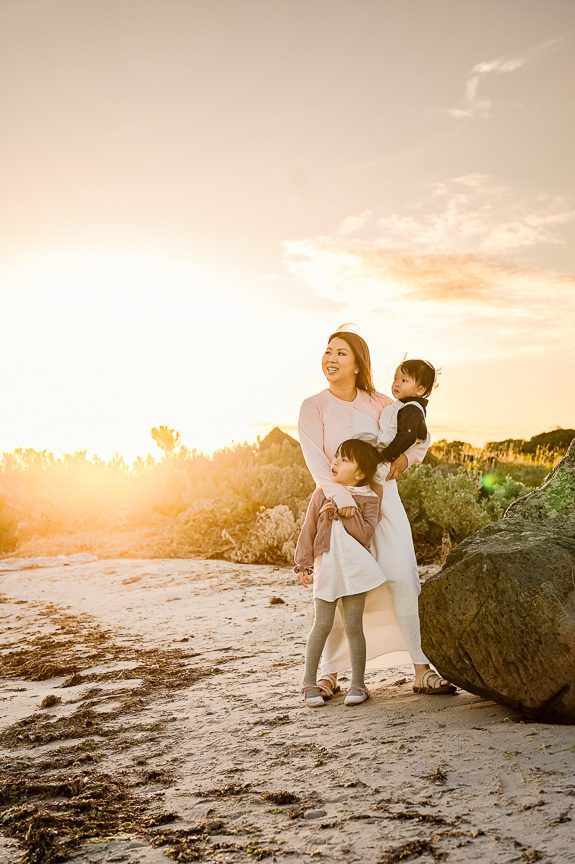 A woman stands on a sandy beach at sunset during one of the best photography session locations, smiling and holding a young child while another stands beside her. Bushes and rocks are visible in the background under a bright sky.